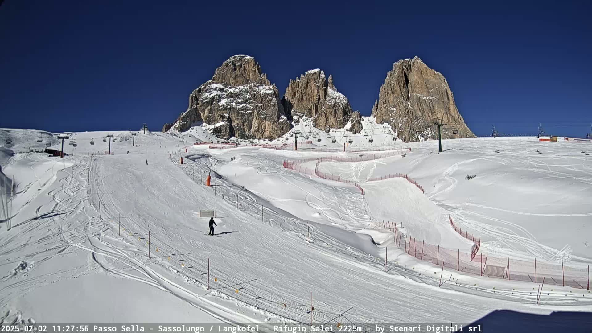 A snowy ski slope with several ski lifts and a few skiers, set against a backdrop of towering, rocky mountains under a clear blue sky.
