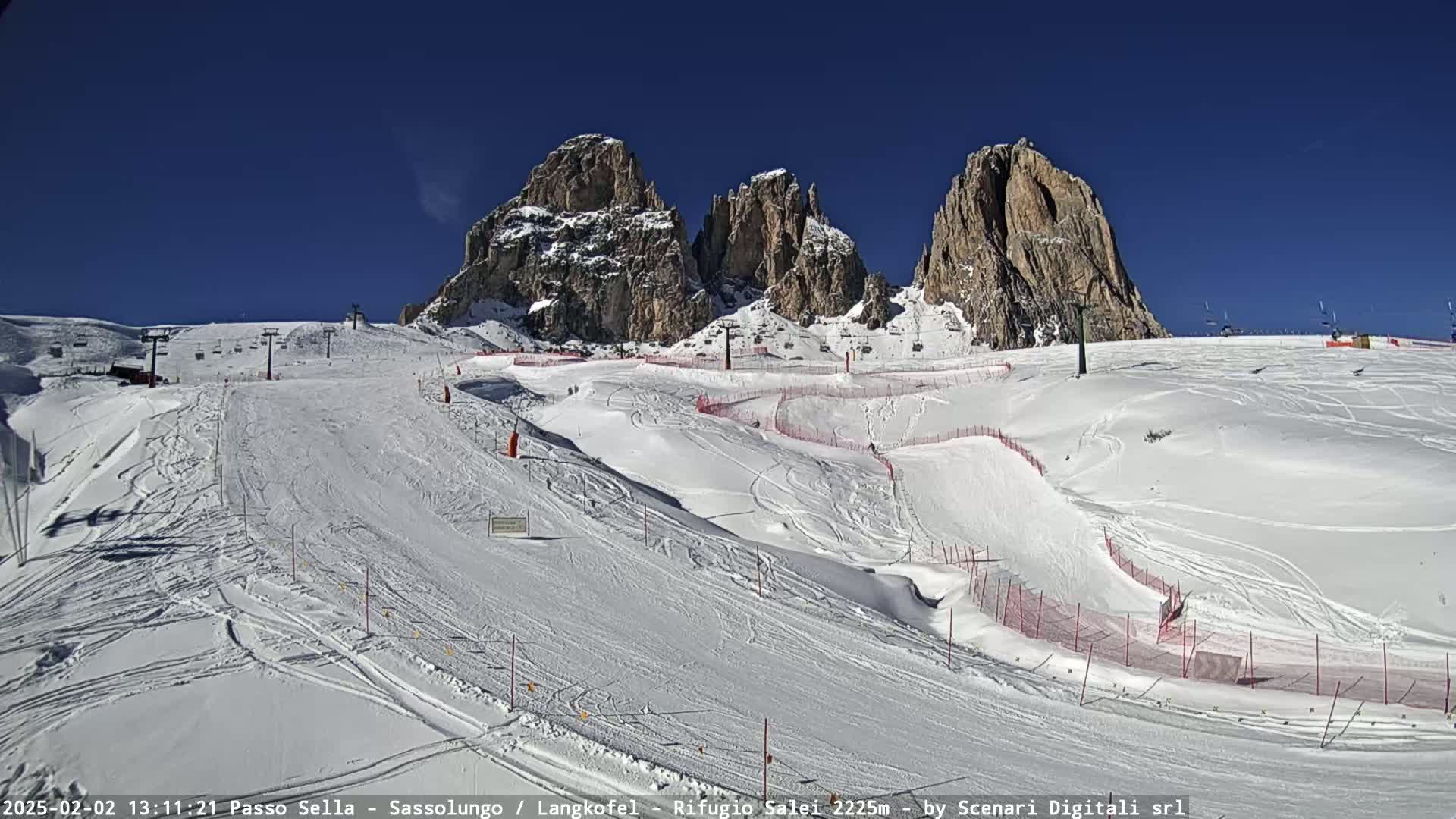A snow-covered ski slope with several ski lifts and marked runs is nestled between tall, rocky mountains under a clear blue sky.