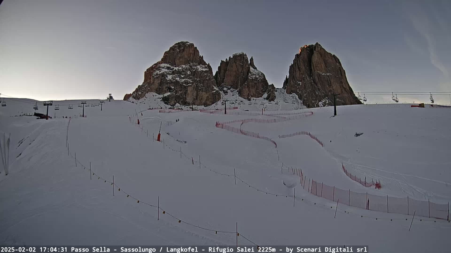 A snow-covered ski slope with chairlifts and fencing winds around towering rock formations under a clear, dusky sky.
