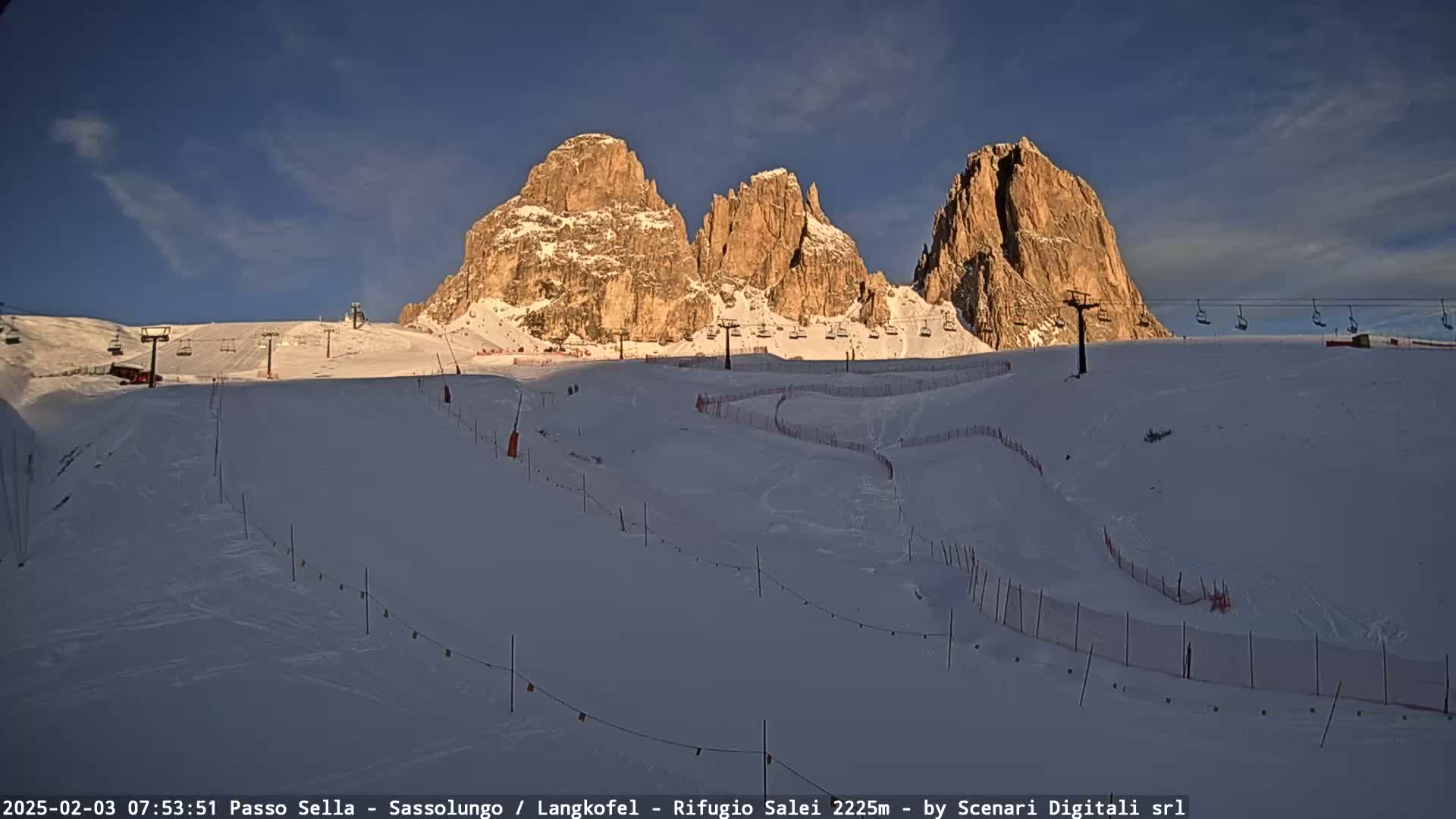 A snow-covered ski slope with chairlifts and tall, rocky mountains in the background under a clear, bright sky.