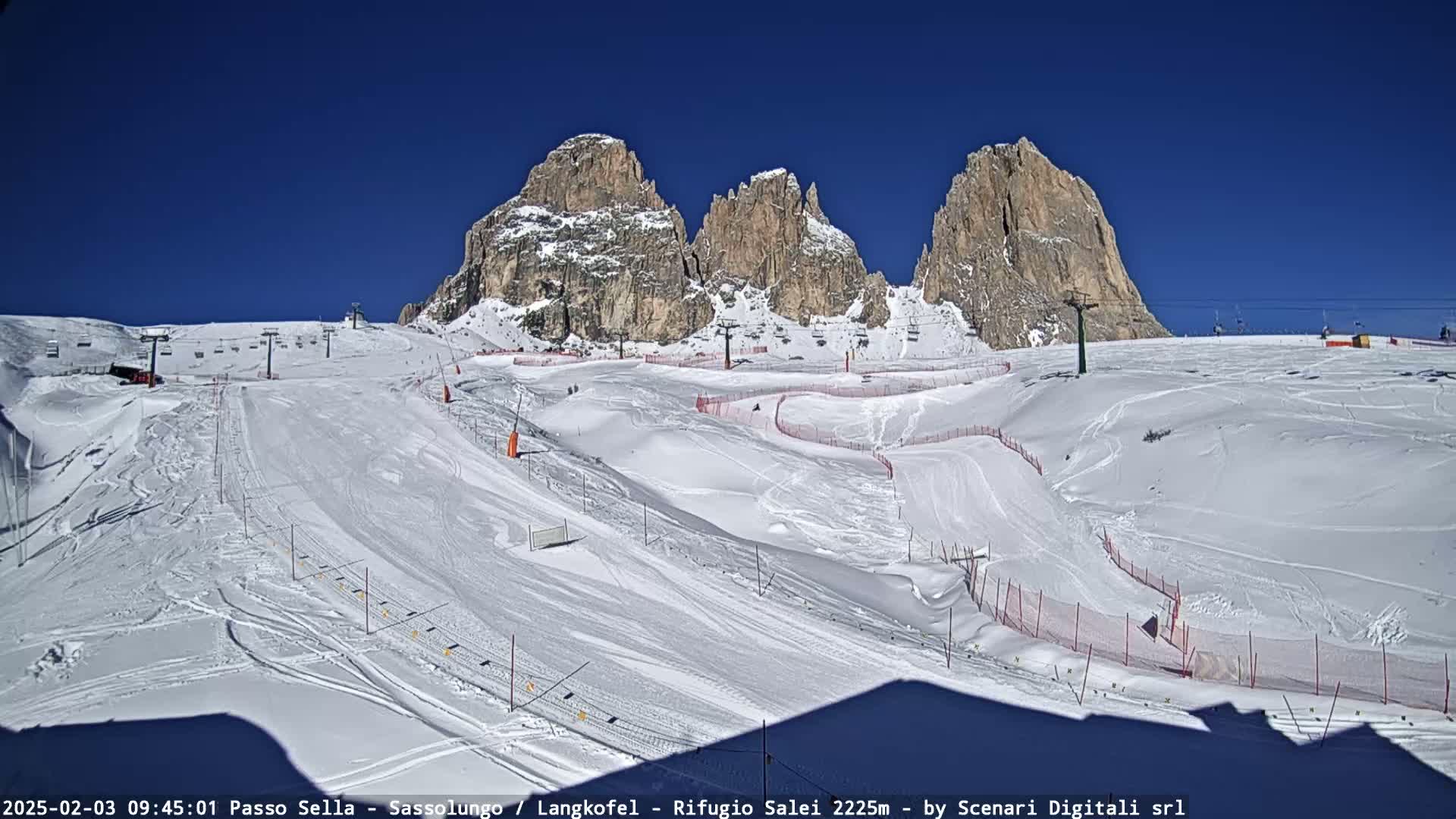 A snow-covered ski slope with ski lifts and marked runs is situated beneath a clear blue sky and imposing rocky mountains.