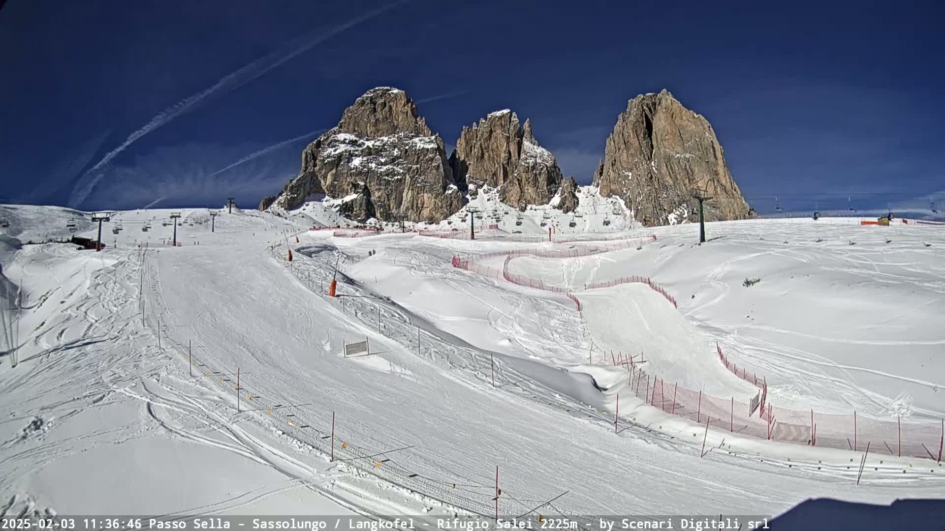A snow-covered ski slope with several ski lifts and marked runs is situated beneath towering, rocky mountains under a clear, sunny sky.
