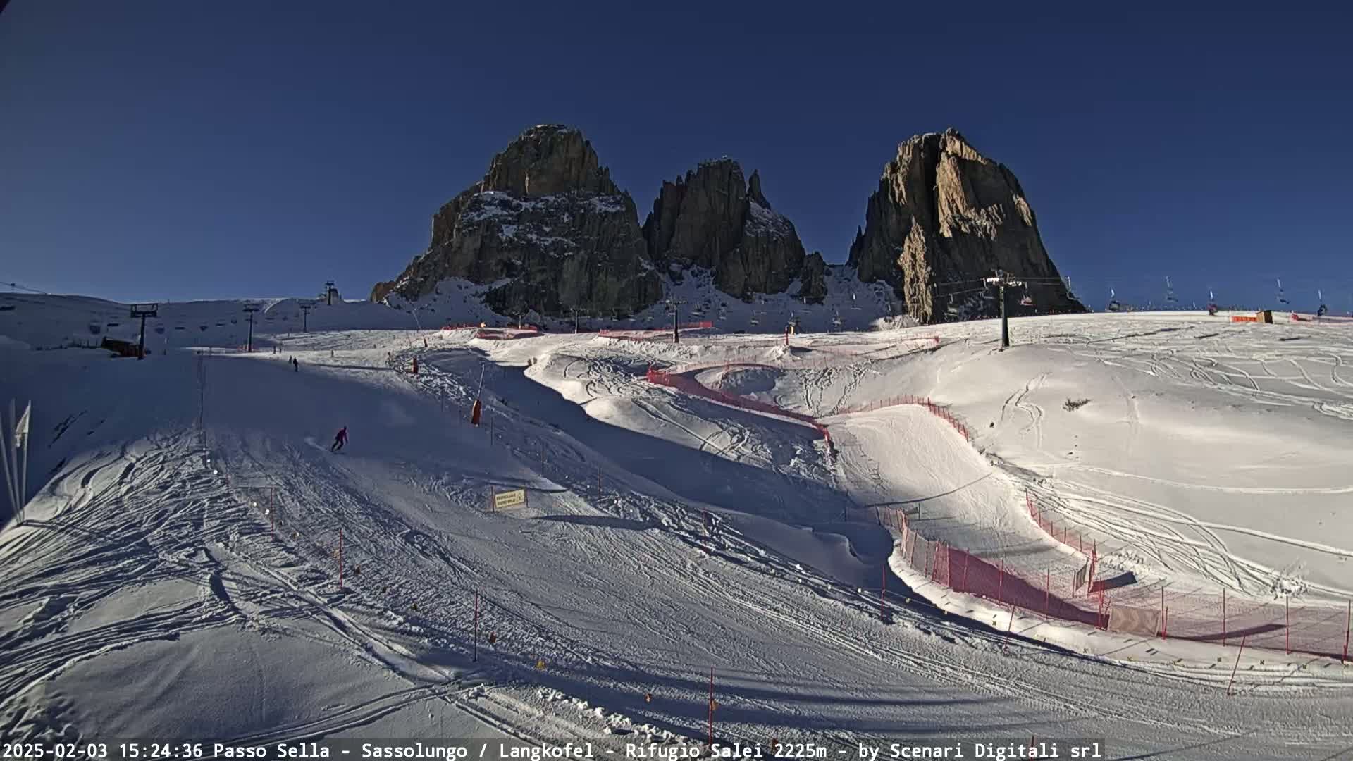 A sunny, snow-covered ski slope with several runs and ski lifts is nestled beneath towering rock formations.
