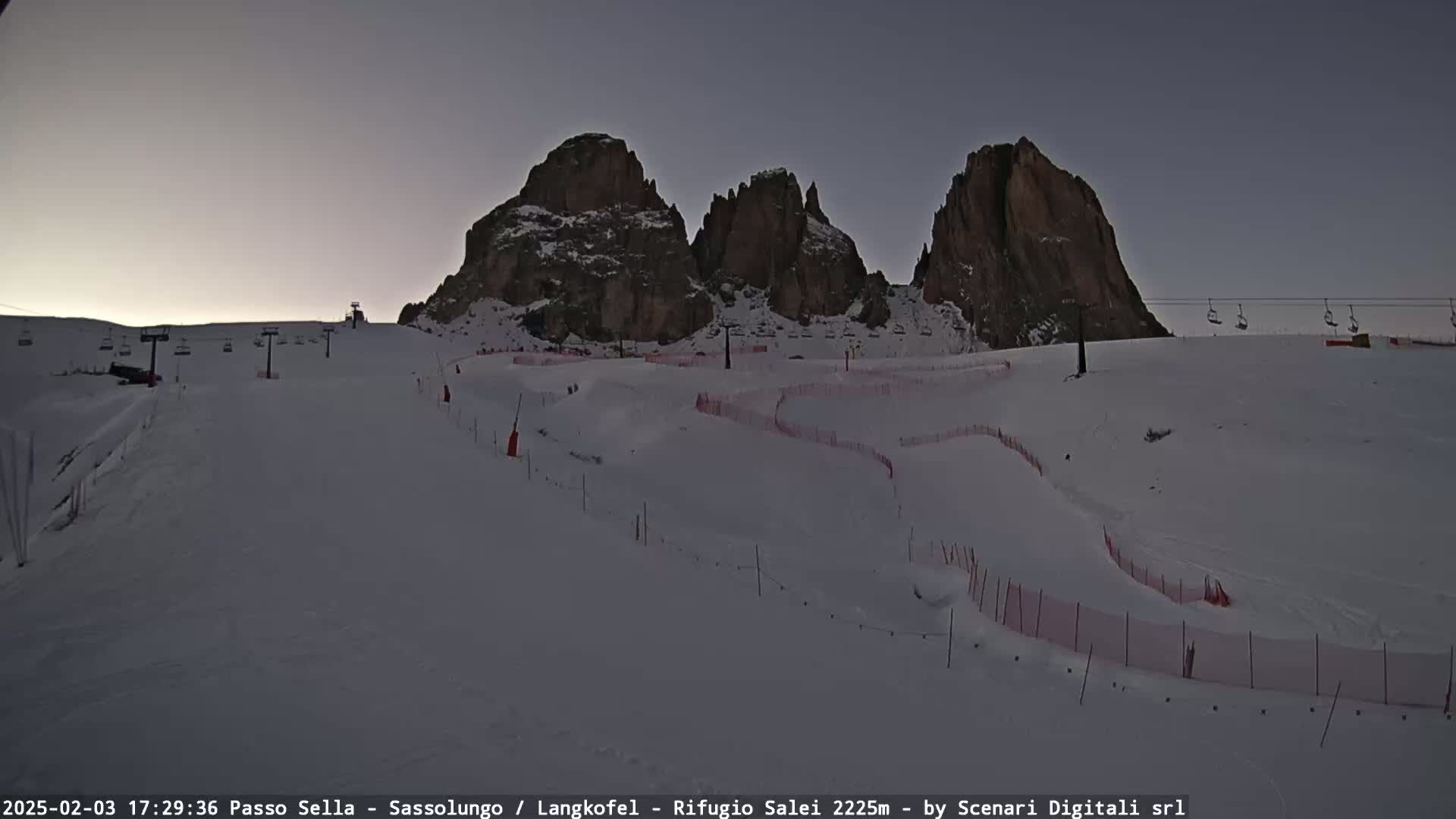 A snowy ski slope with chairlifts and red fencing winds below a range of dark, rocky mountains under a twilight sky.
