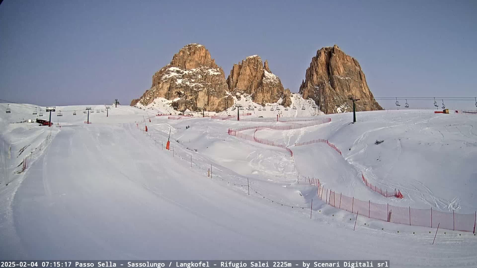 A snow-covered ski slope with several chairlifts and orange safety netting winds around the base of three rocky peaks under a clear blue sky.