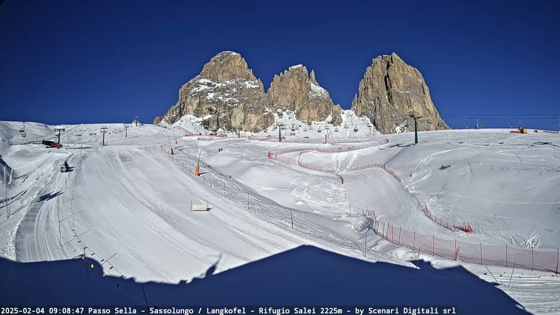 A snow-covered ski slope with several ski lifts and marked runs is situated beneath a clear blue sky and imposing, rocky mountains.