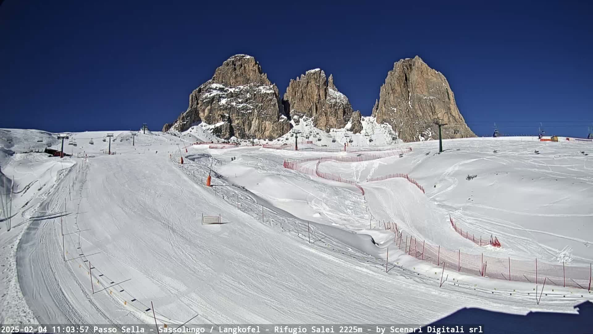 A snow-covered ski slope with marked runs and lift infrastructure is situated below a clear blue sky and imposing rocky mountain peaks.