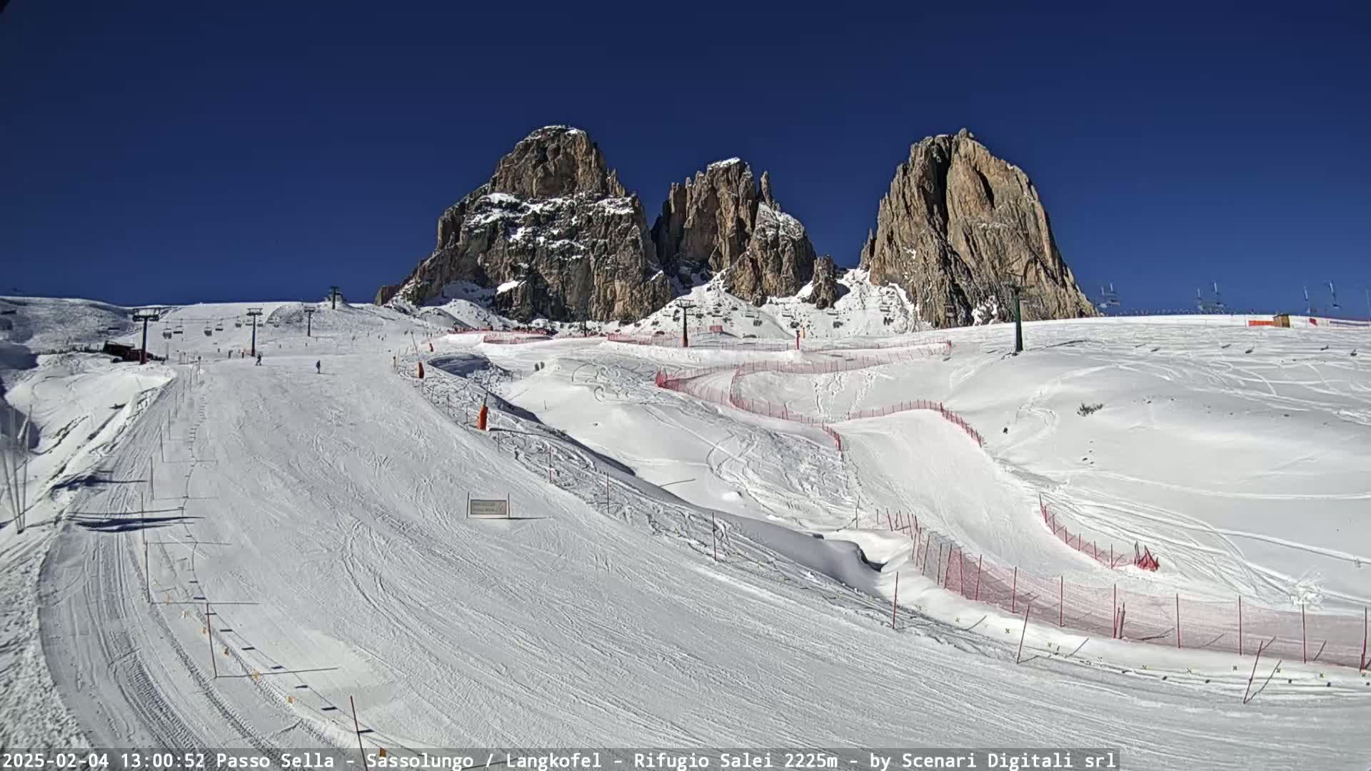 A sunny, snow-covered ski slope with several ski runs and a chairlift, set against a backdrop of towering, snow-dusted mountains.