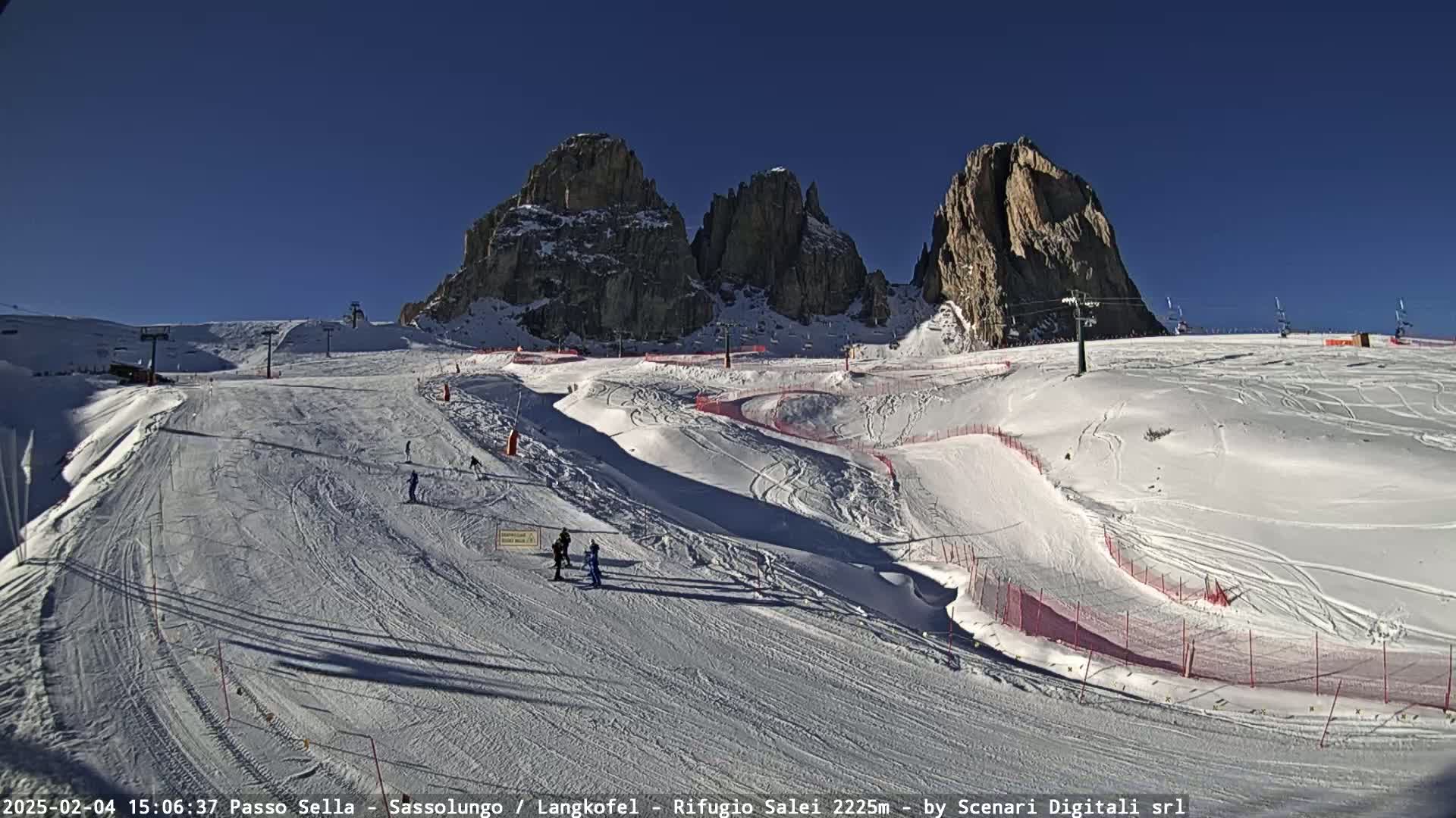 A sunny day on a snow-covered ski slope in the mountains, with several skiers visible and towering rock formations in the background.