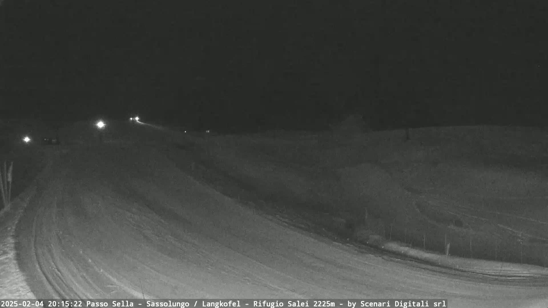 A nighttime, snow-covered road curves gently to the right with a few vehicles visible in the distance under dark skies.
