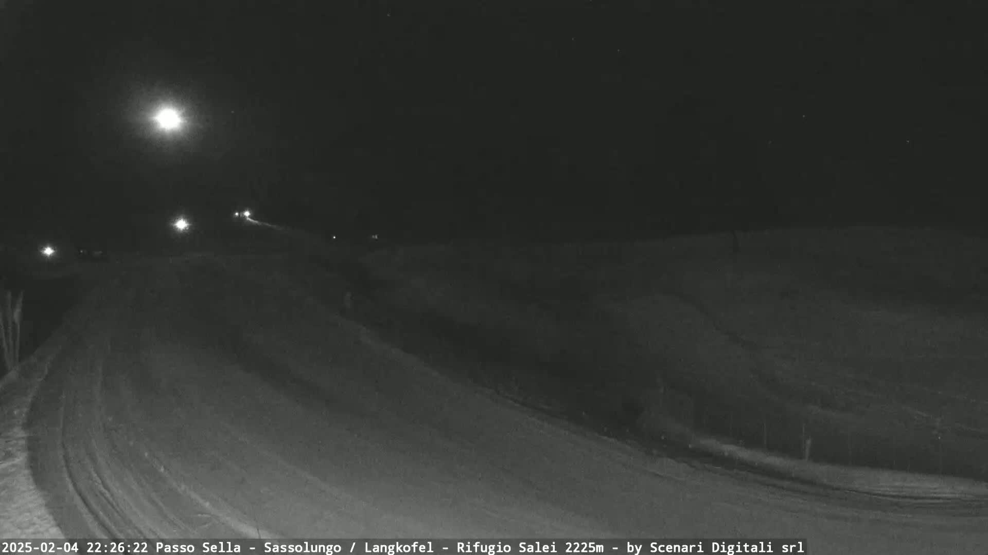 A snow-covered road curves gently under a bright moon at night, with a few vehicle headlights visible in the distance.