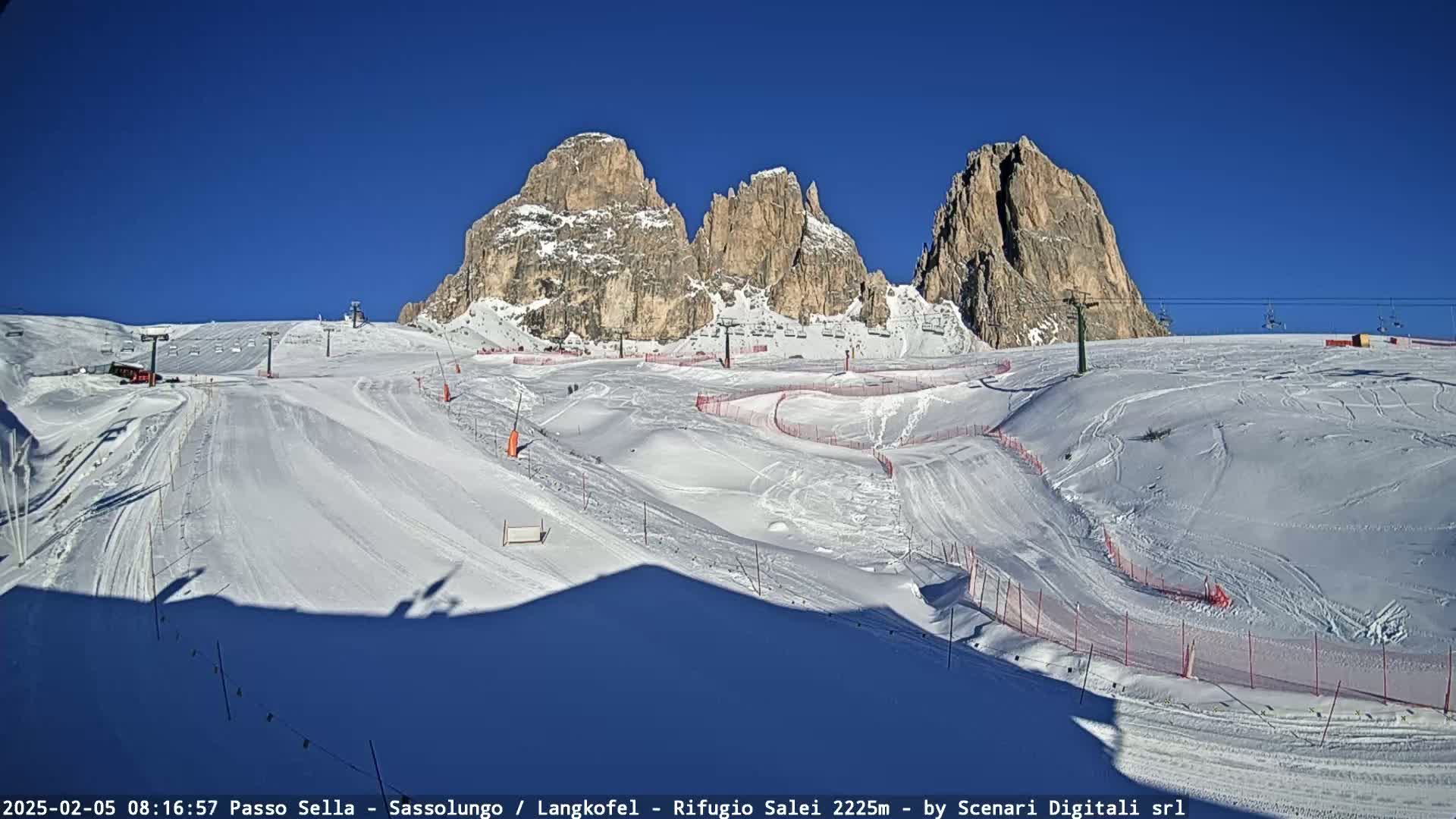 A snow-covered ski slope with ski lifts and several marked runs is situated below a range of rugged, snow-dusted mountains under a clear blue sky.