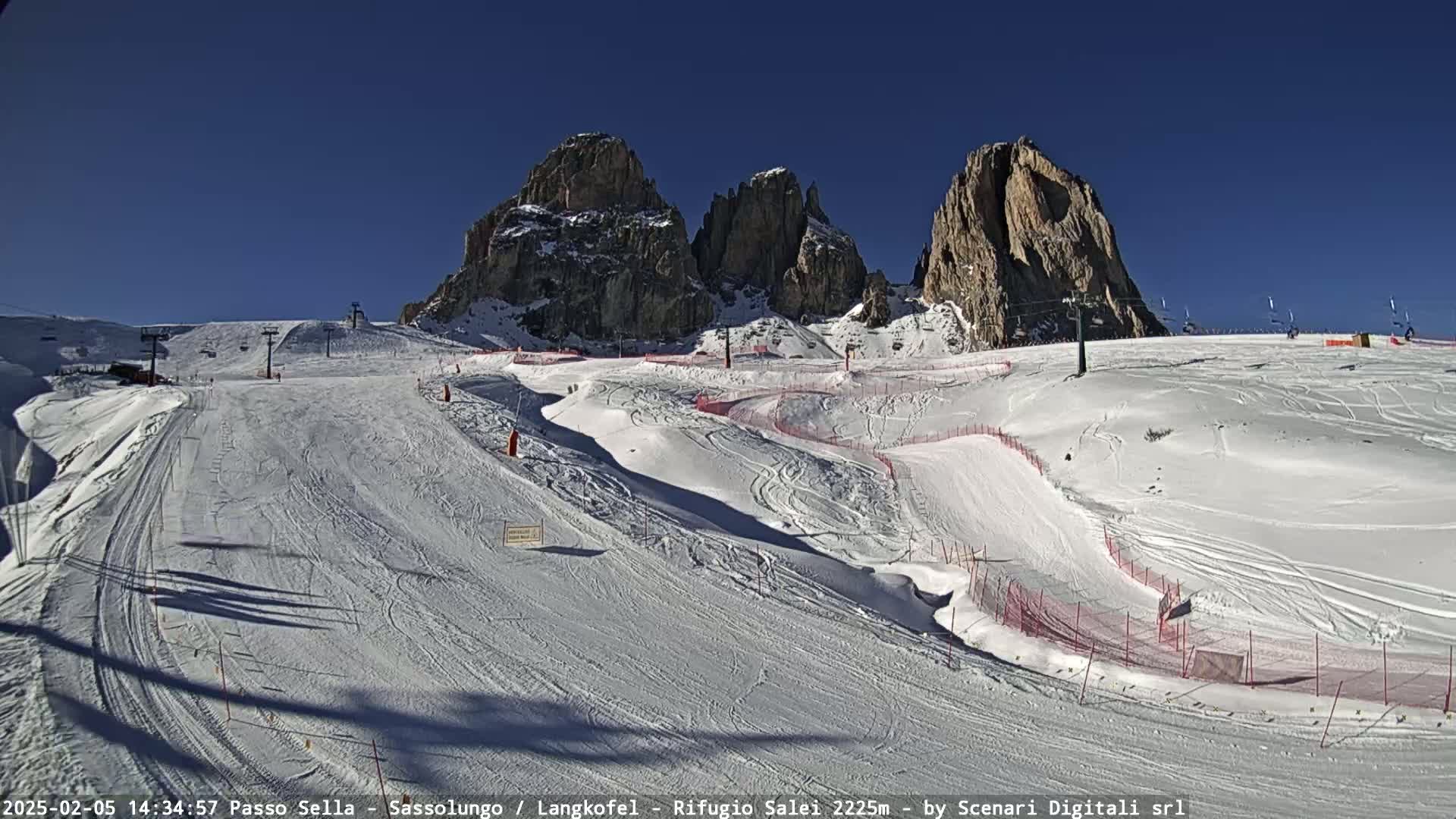 A snow-covered ski slope with several runs and ski lifts is situated below rocky, snow-dusted peaks under a clear blue sky.