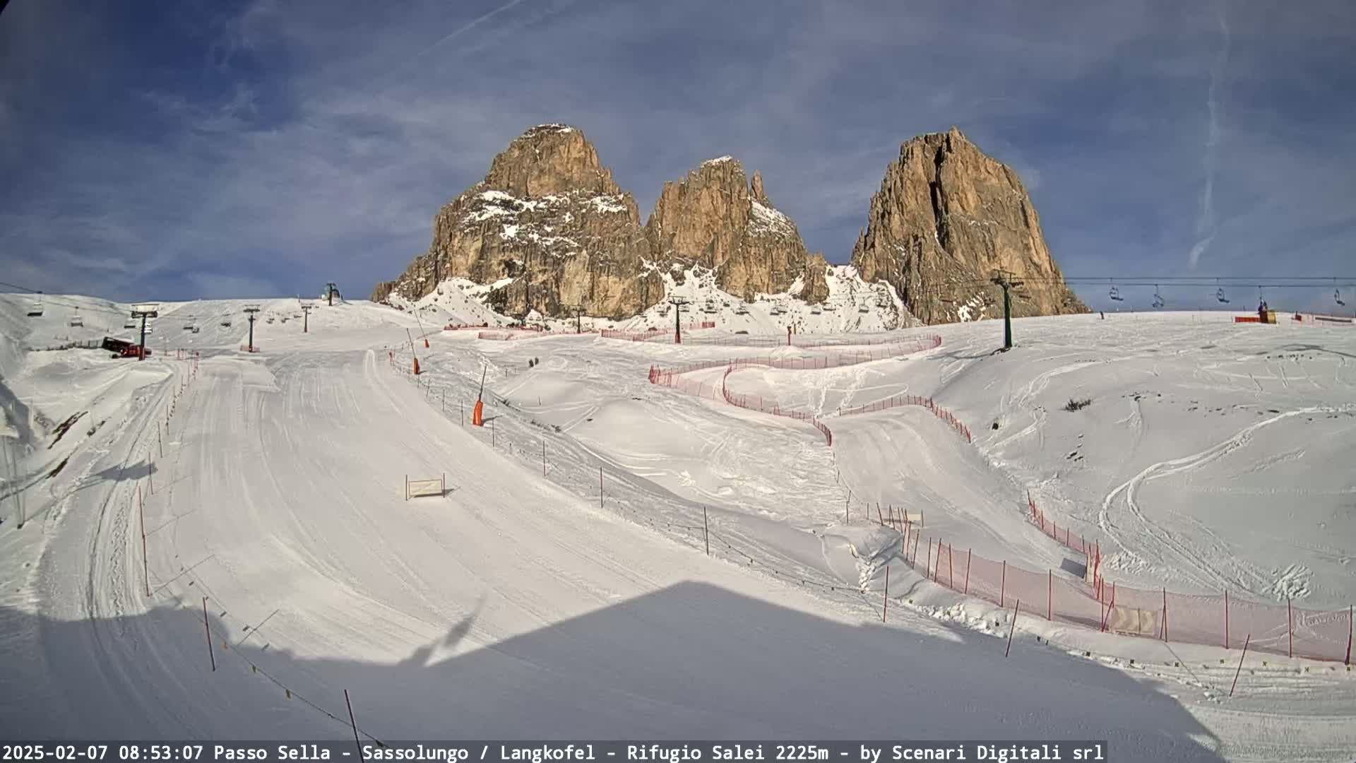 A snow-covered ski slope with chairlifts and several rocky peaks in the background under a mostly sunny sky.