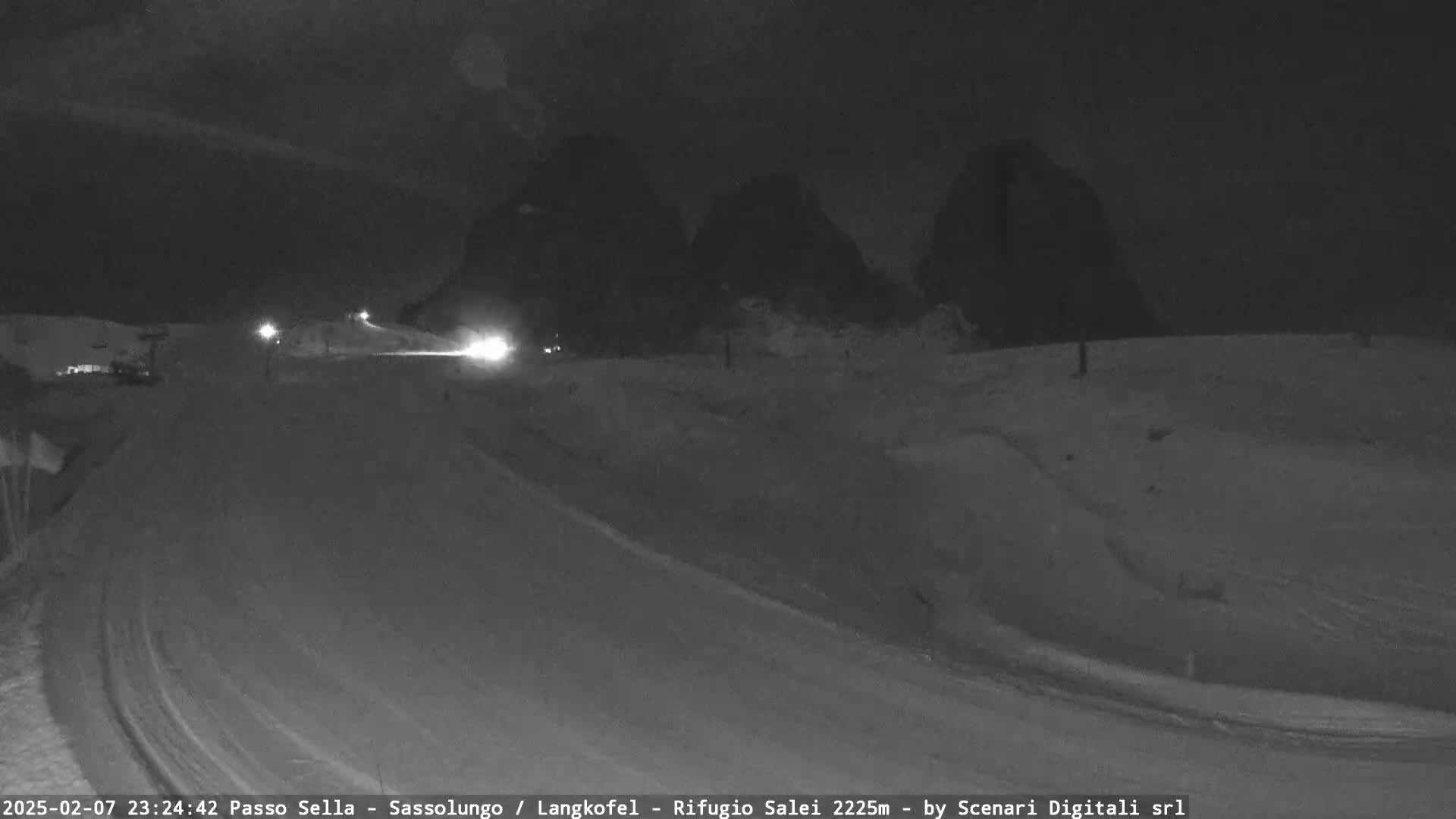 A nighttime, snow-covered road curves around a mountain range under a dark sky.