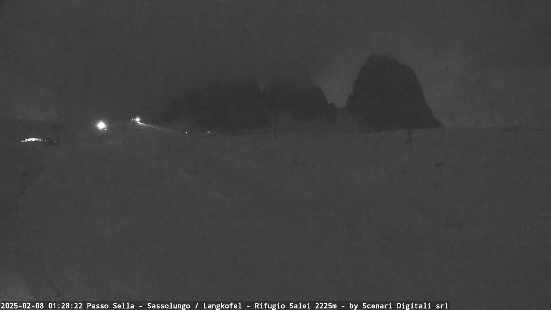 A nighttime, snowy scene shows a road with headlights visible in the distance, and dark, snow-covered land with indistinct shapes in the background.
