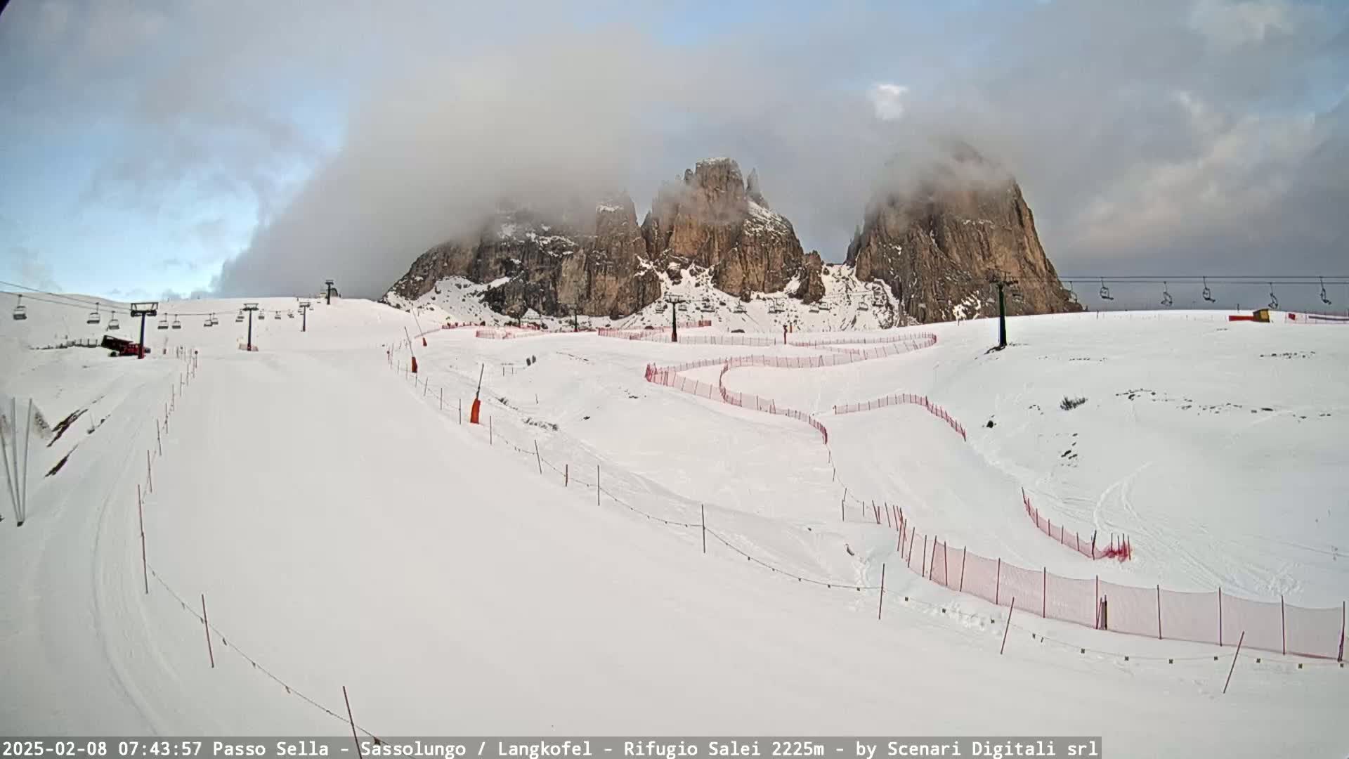 A snowy ski slope with a chairlift and red safety netting is situated beneath partially cloud-covered rocky mountains.