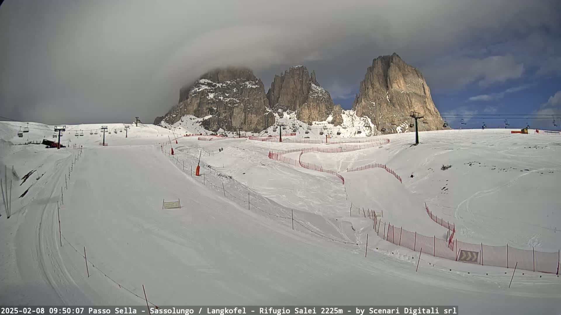 A snow-covered ski slope with ski lifts and mountain peaks in the background under a partly cloudy sky.