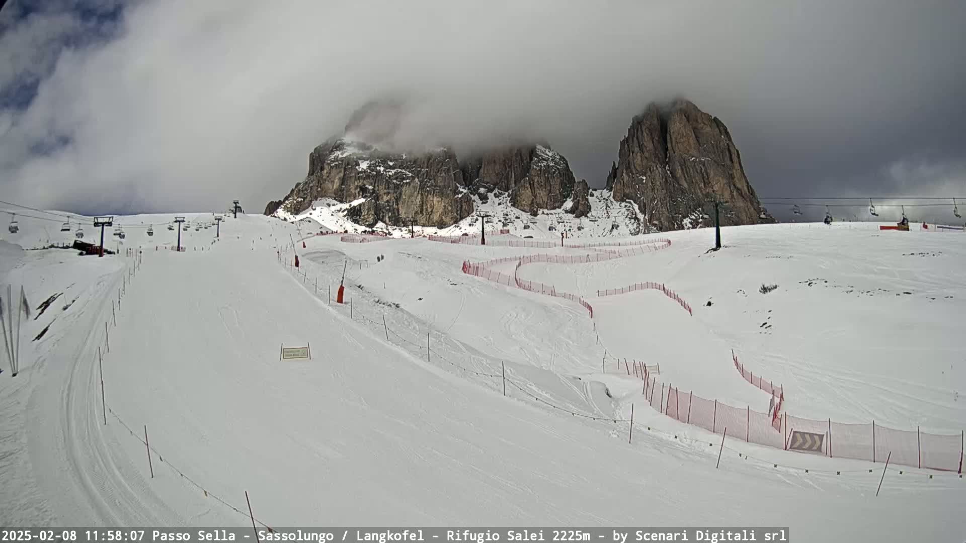 A snow-covered ski slope with a chairlift and red safety netting, partially obscured by clouds, is situated in front of rocky mountain peaks.