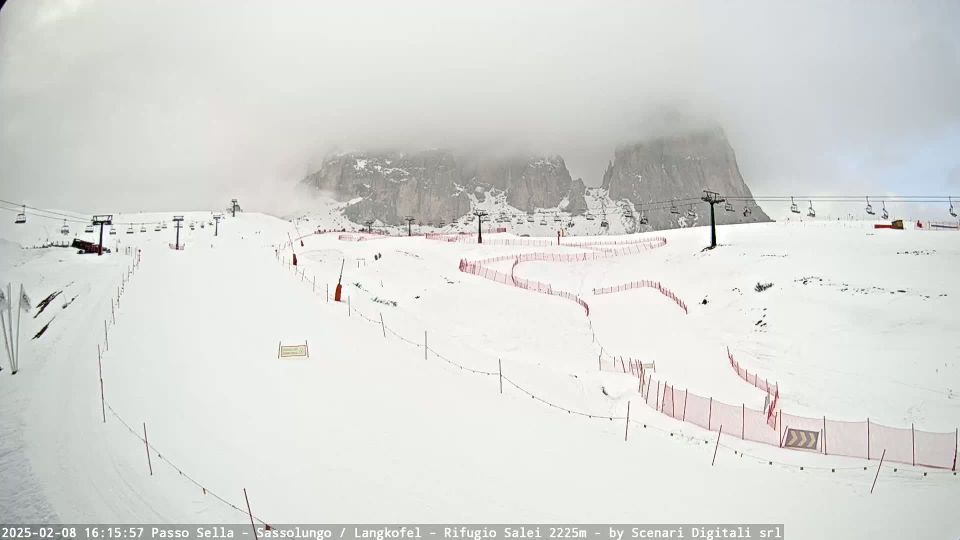 A snowy ski slope with chairlifts and red safety nets is visible beneath a foggy sky, mountains looming in the background.