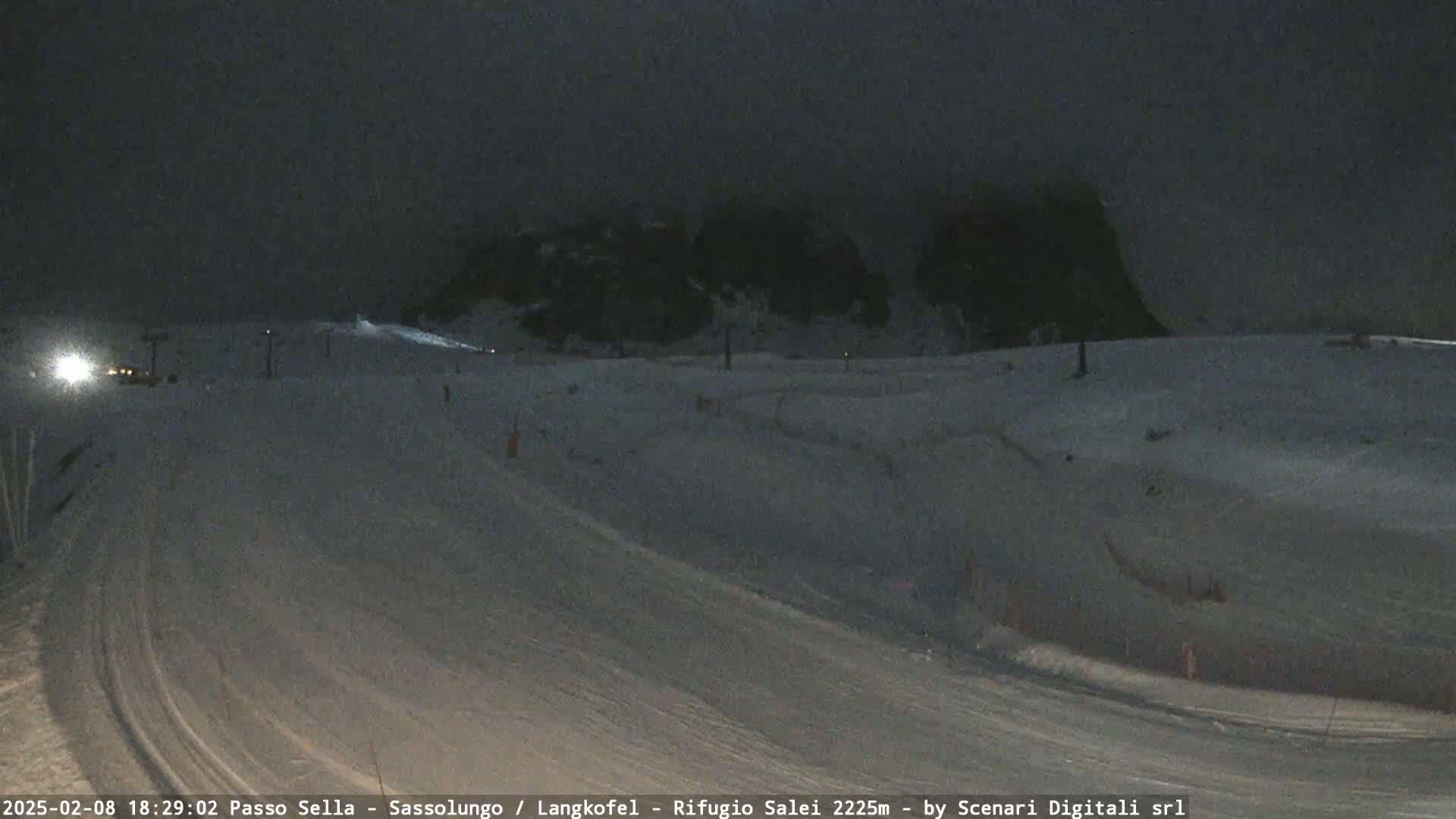A snow-covered ski slope at night, with dark, cloudy skies and dimly lit mountains in the background.