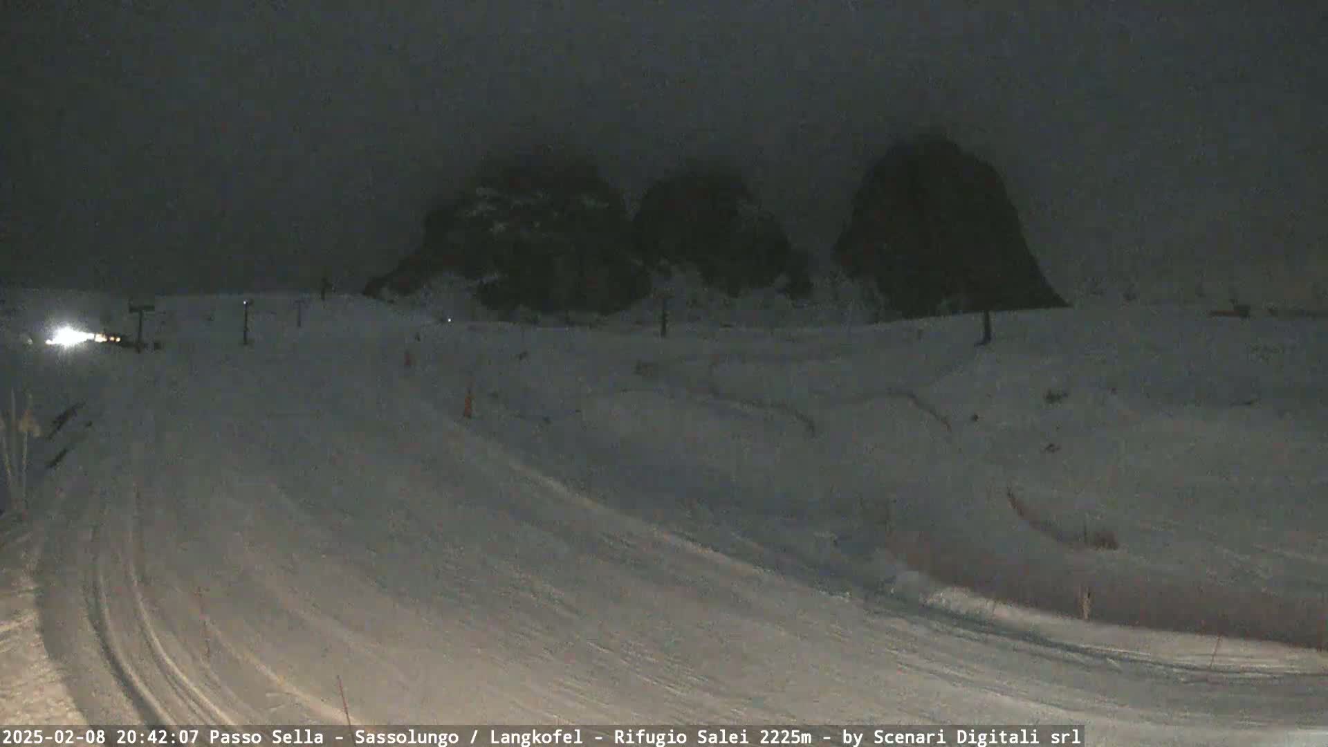 A snow-covered ski slope at night, illuminated by a single headlight, with dark mountain silhouettes in the background under a cloudy sky.