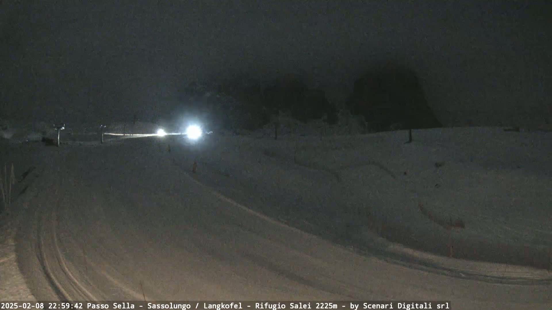 A nighttime snow-covered road with vehicle headlights visible in the distance, under a dark, cloudy sky.