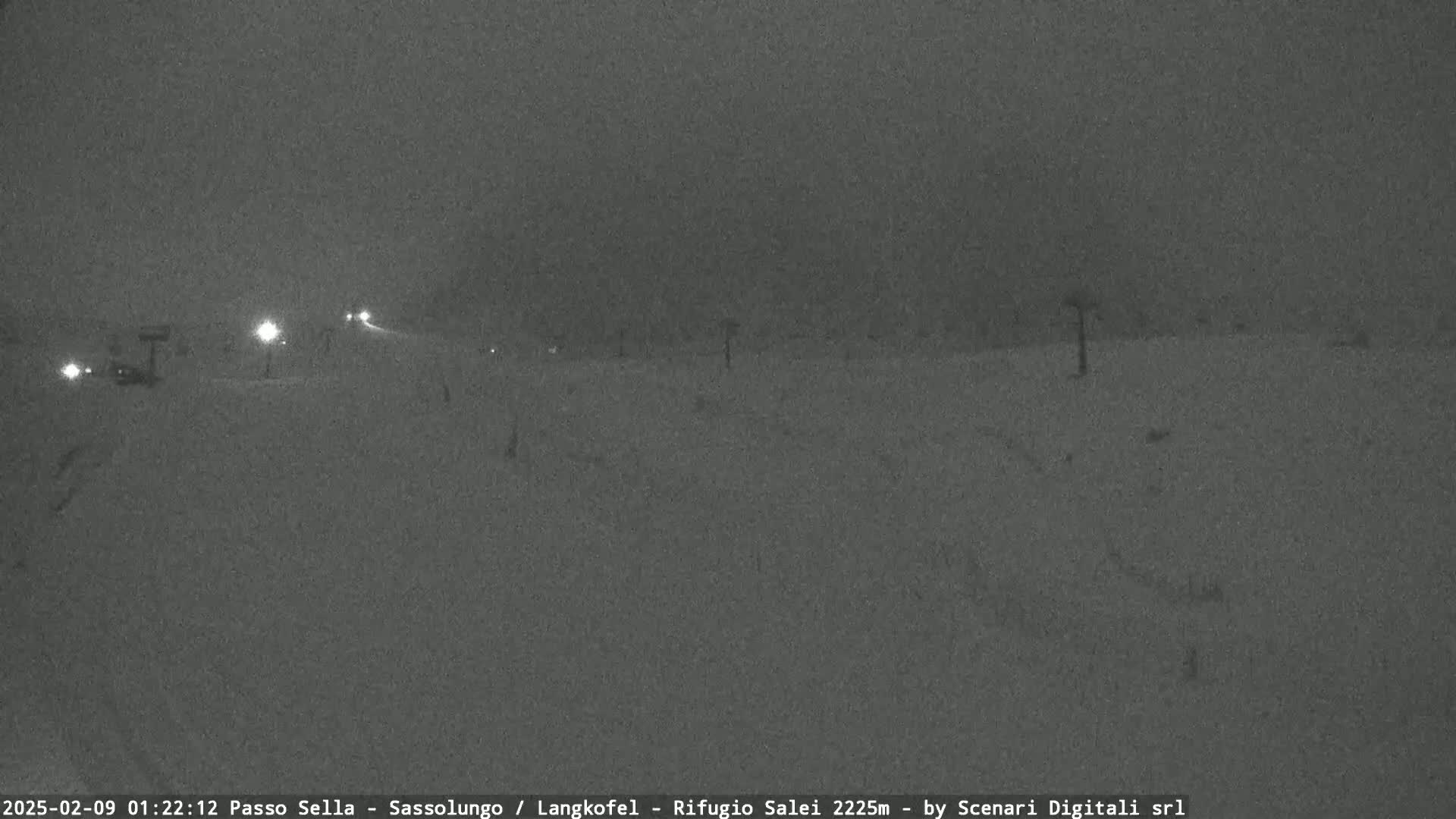A nighttime, snow-covered road scene is dimly lit by vehicle headlights and streetlights in snowy conditions.