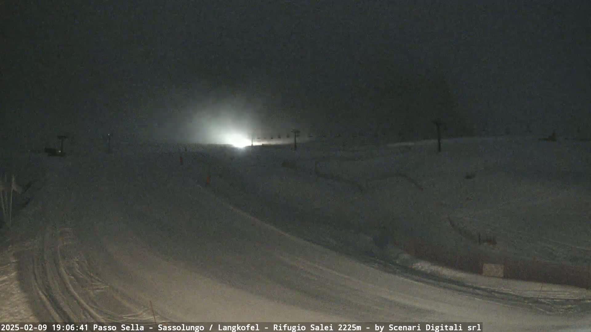 A snow-covered slope at night is lightly illuminated by a distant light source in snowy conditions.