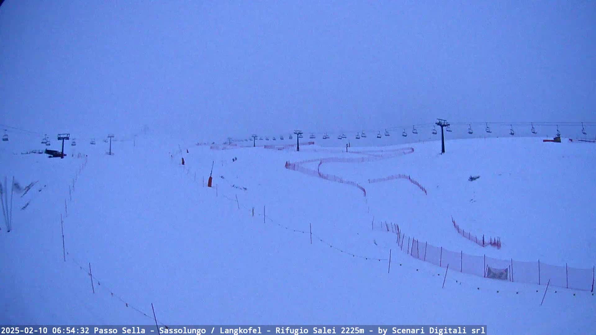 A snow-covered ski slope with several chairlifts and red safety netting under an overcast sky.