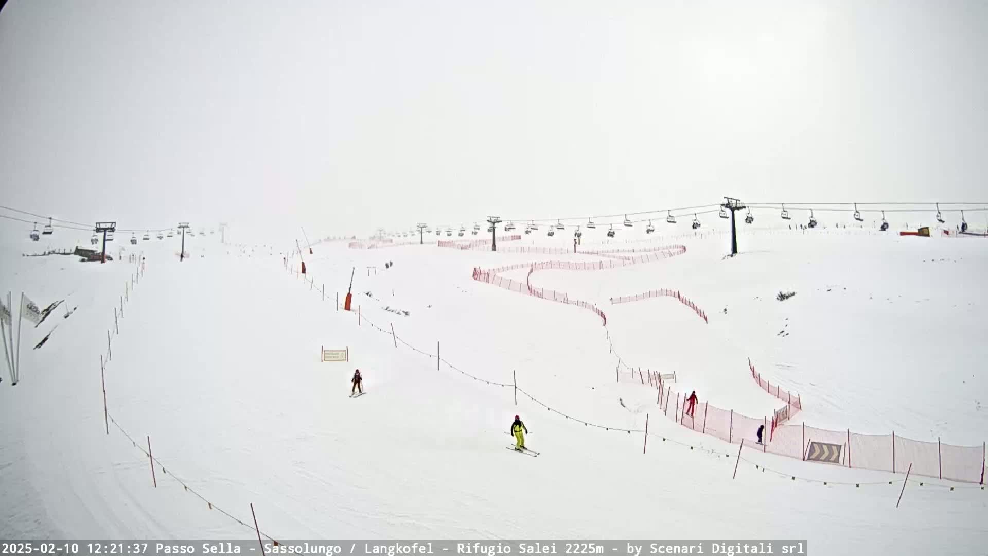 A snowy ski slope with several skiers and chairlifts visible under overcast conditions.