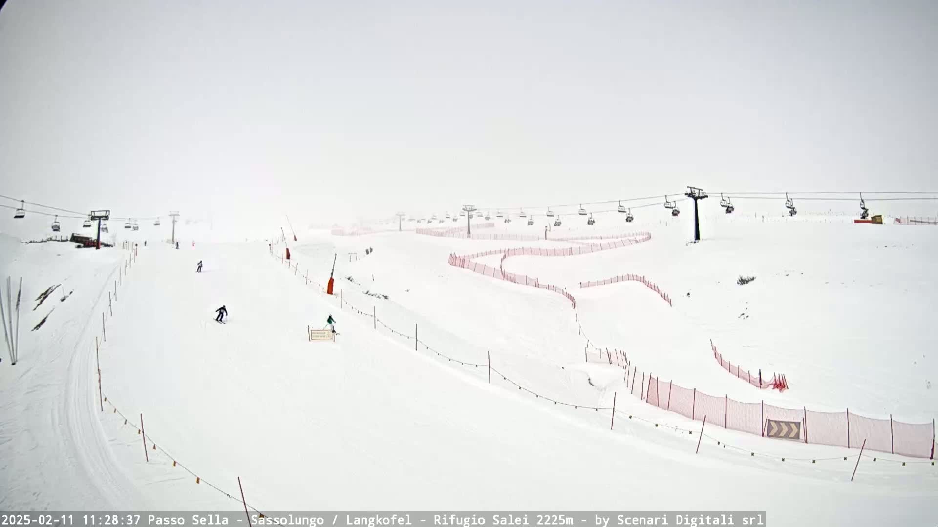 A snowy ski slope with several skiers and chairlifts visible under hazy, overcast conditions.