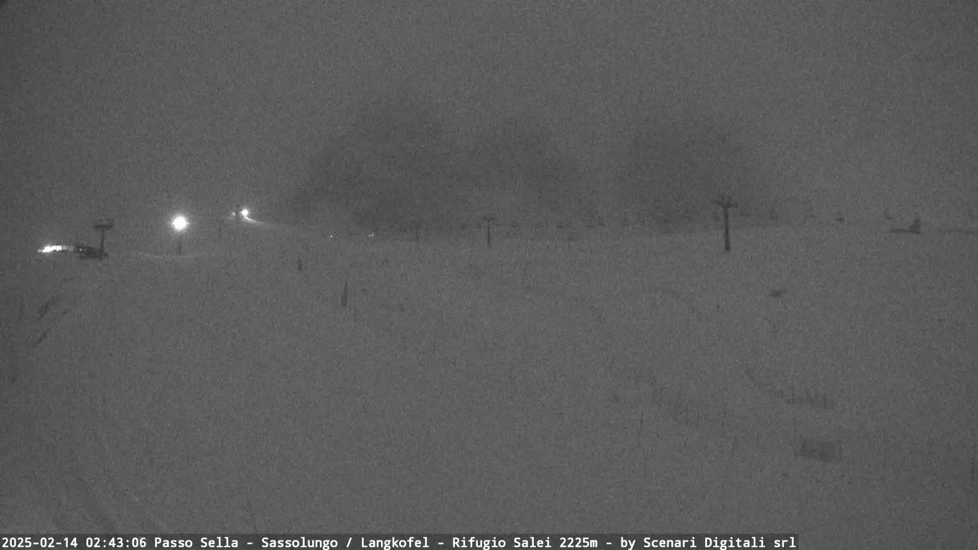 A snow-covered landscape at night is dimly lit by a few lights, with some indistinct objects in the distance under snowy, dark conditions.
