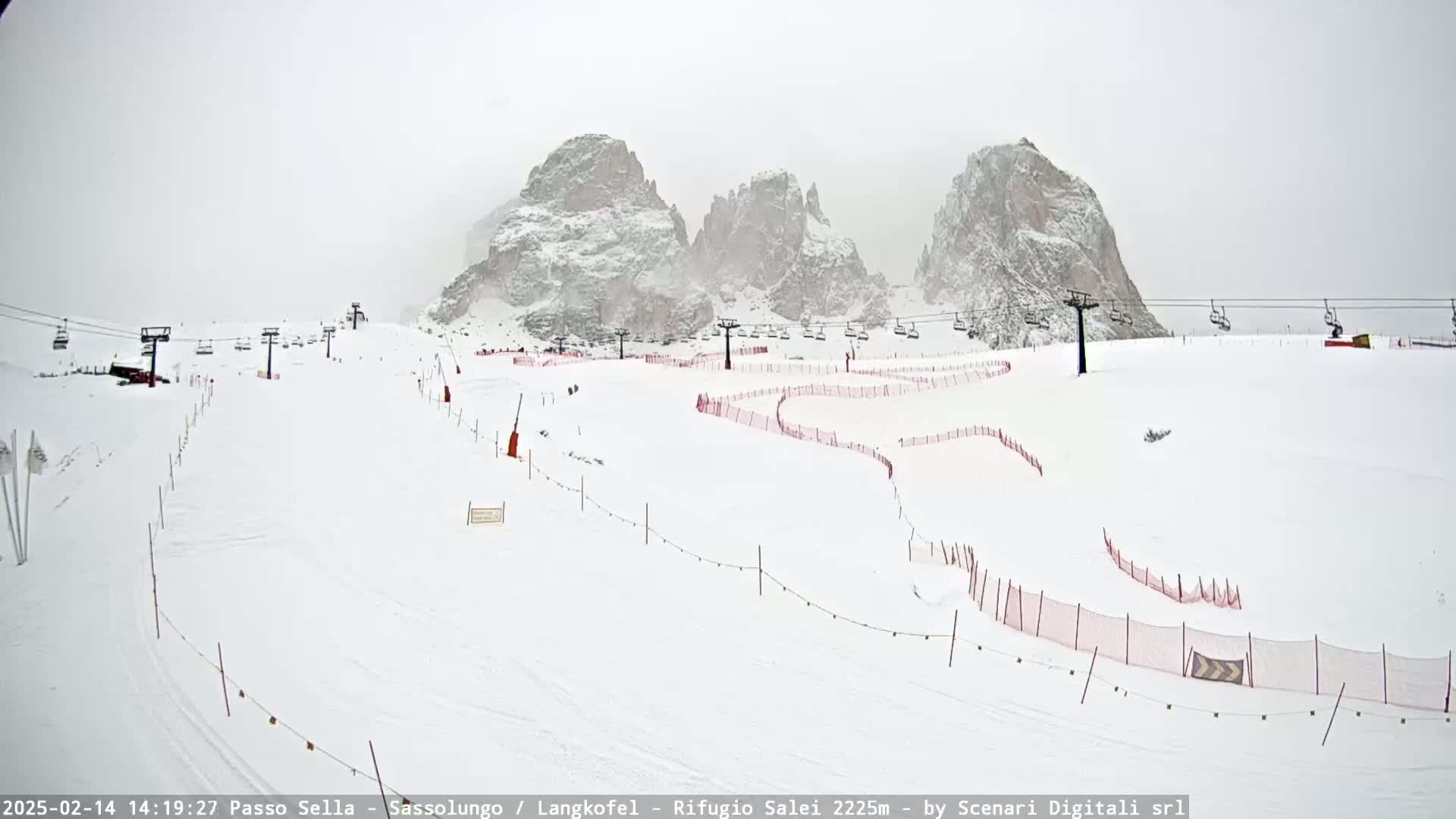 A snowy ski slope with chairlifts and snow-covered mountains in the background under overcast skies.
