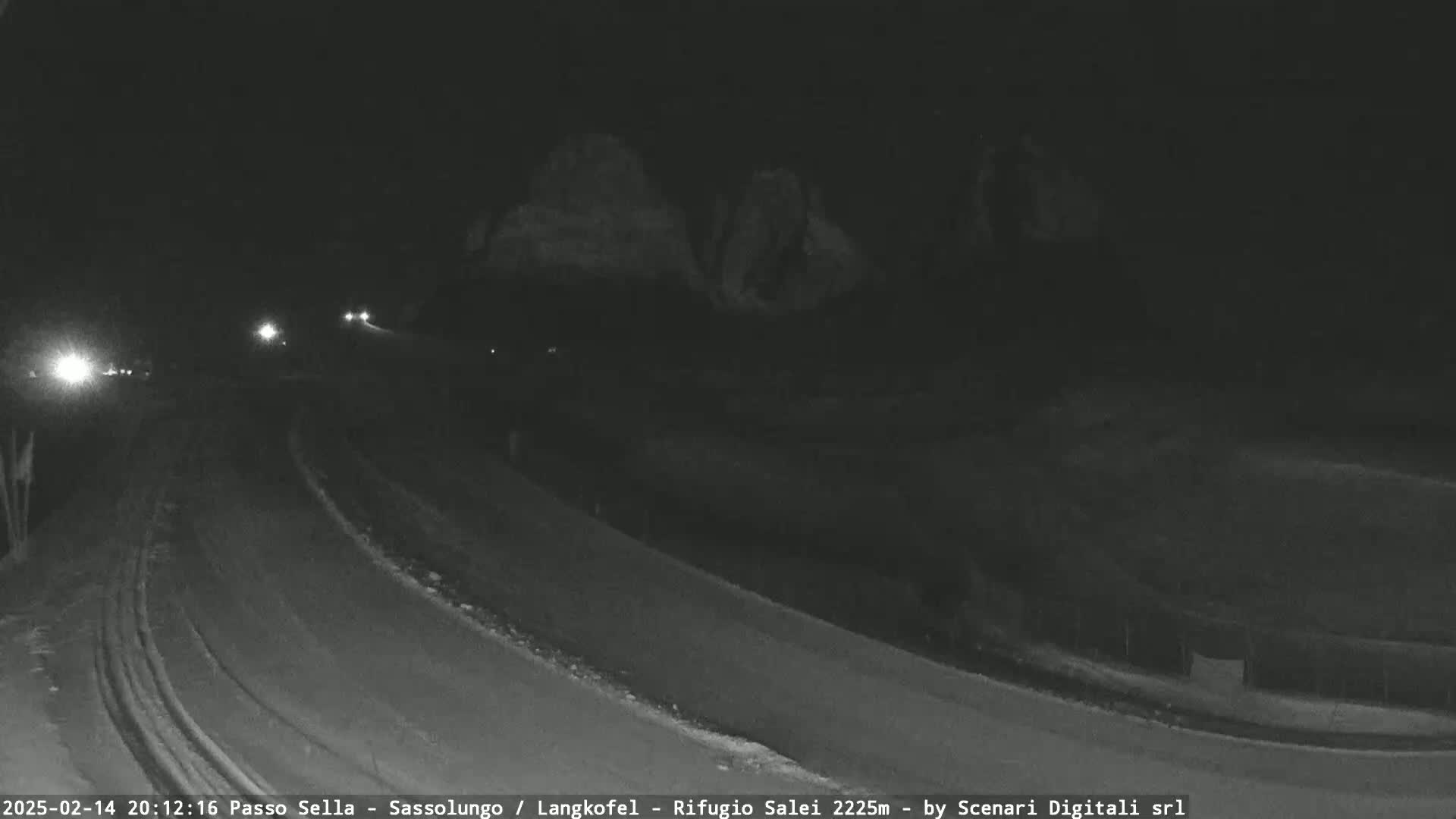 A snow-covered road curves gently at night, with a few distant headlights visible and dark, indistinct shapes of land in the background.