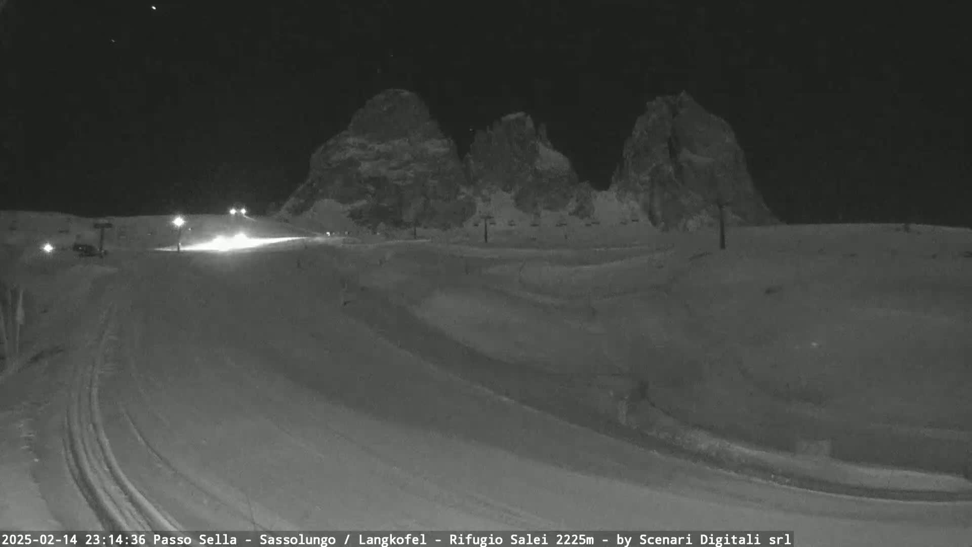 A snow-covered landscape at night is illuminated by several lights, with large, dark mountains in the background.