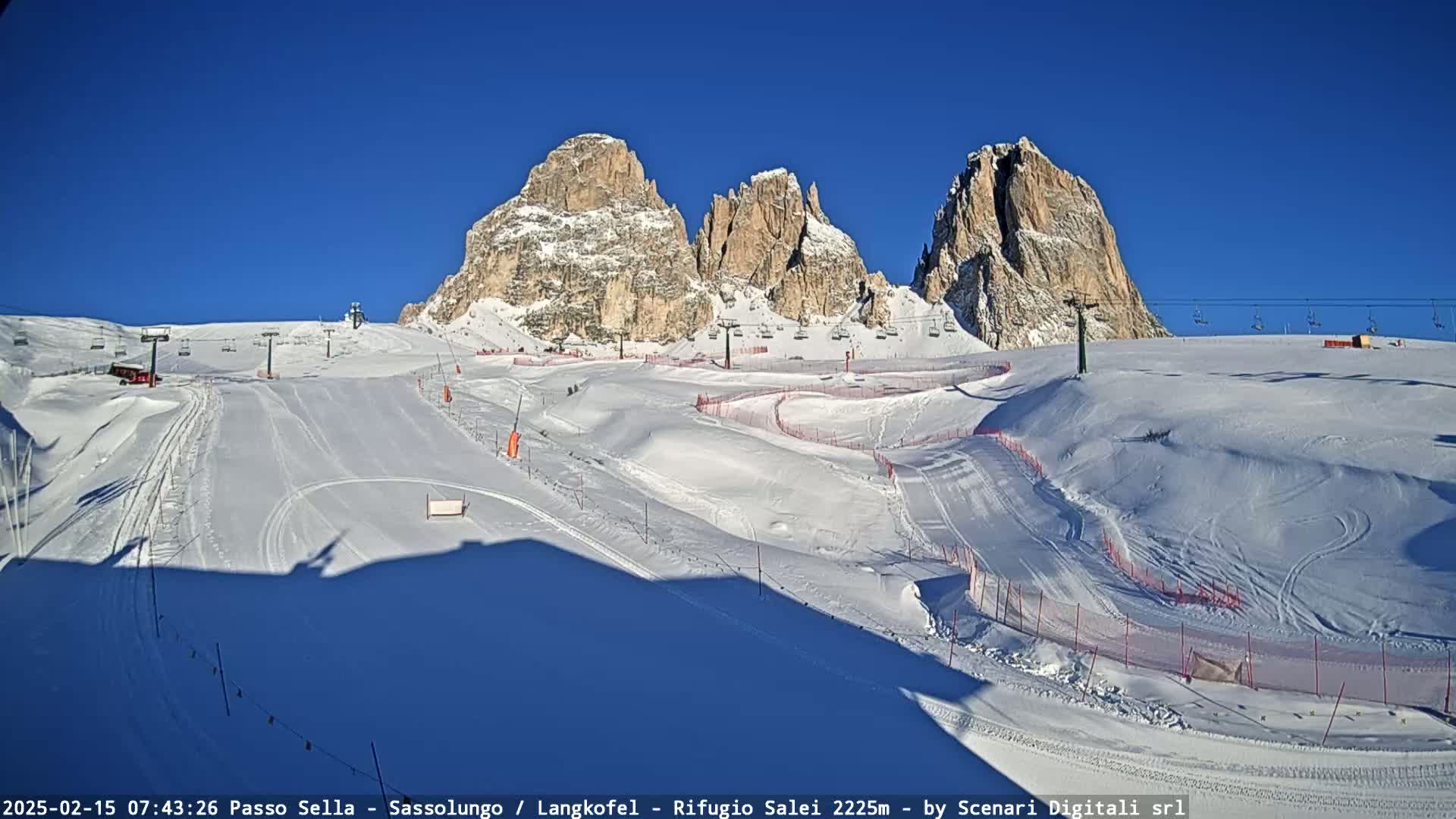 A snow-covered ski slope with several ski lifts and tall, rocky mountains in the background under a clear blue sky.