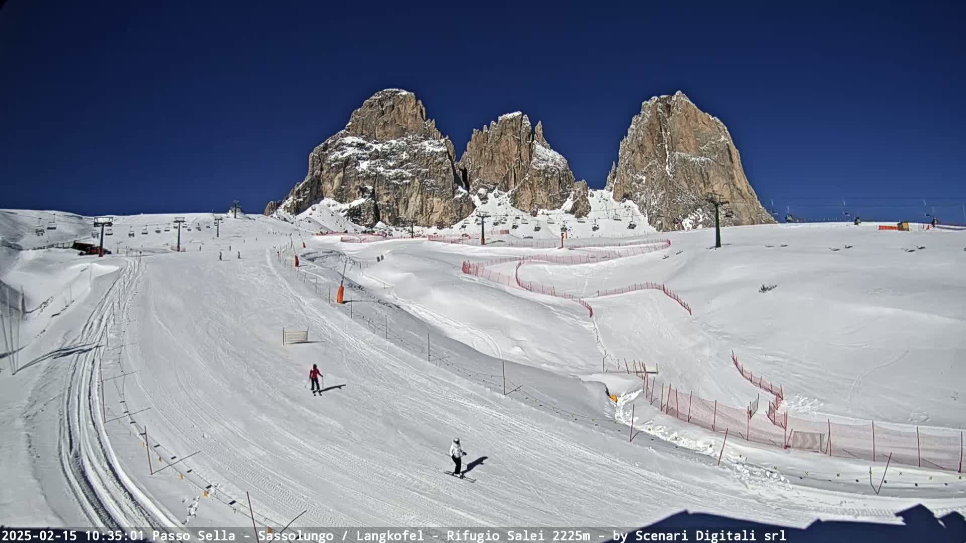 A sunny day at a snow-covered ski resort with several skiers visible on the slopes, situated against a backdrop of towering, rocky mountains.