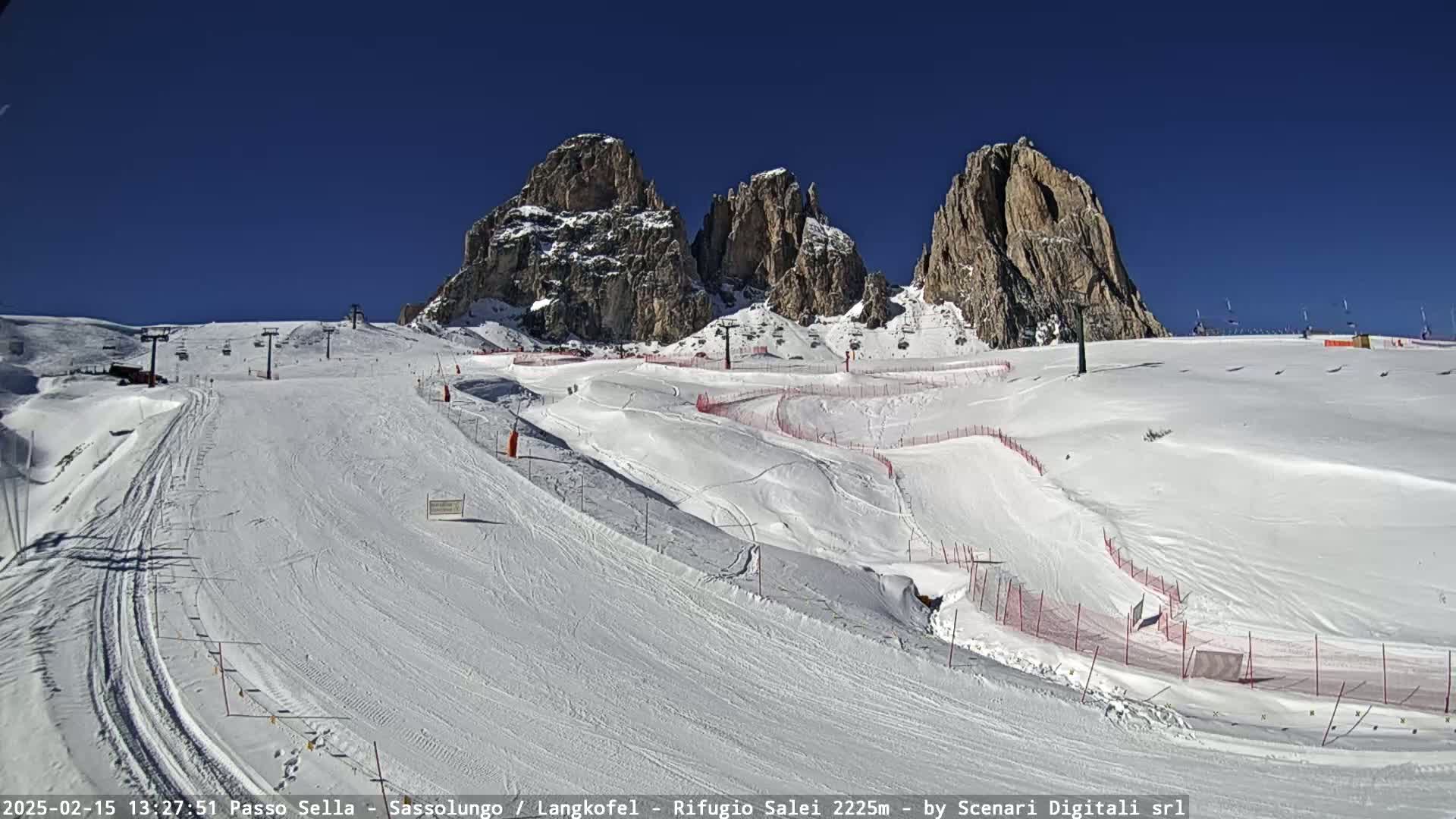 A snow-covered ski slope with several runs and lifts is situated beneath a clear blue sky and towering rocky mountains.