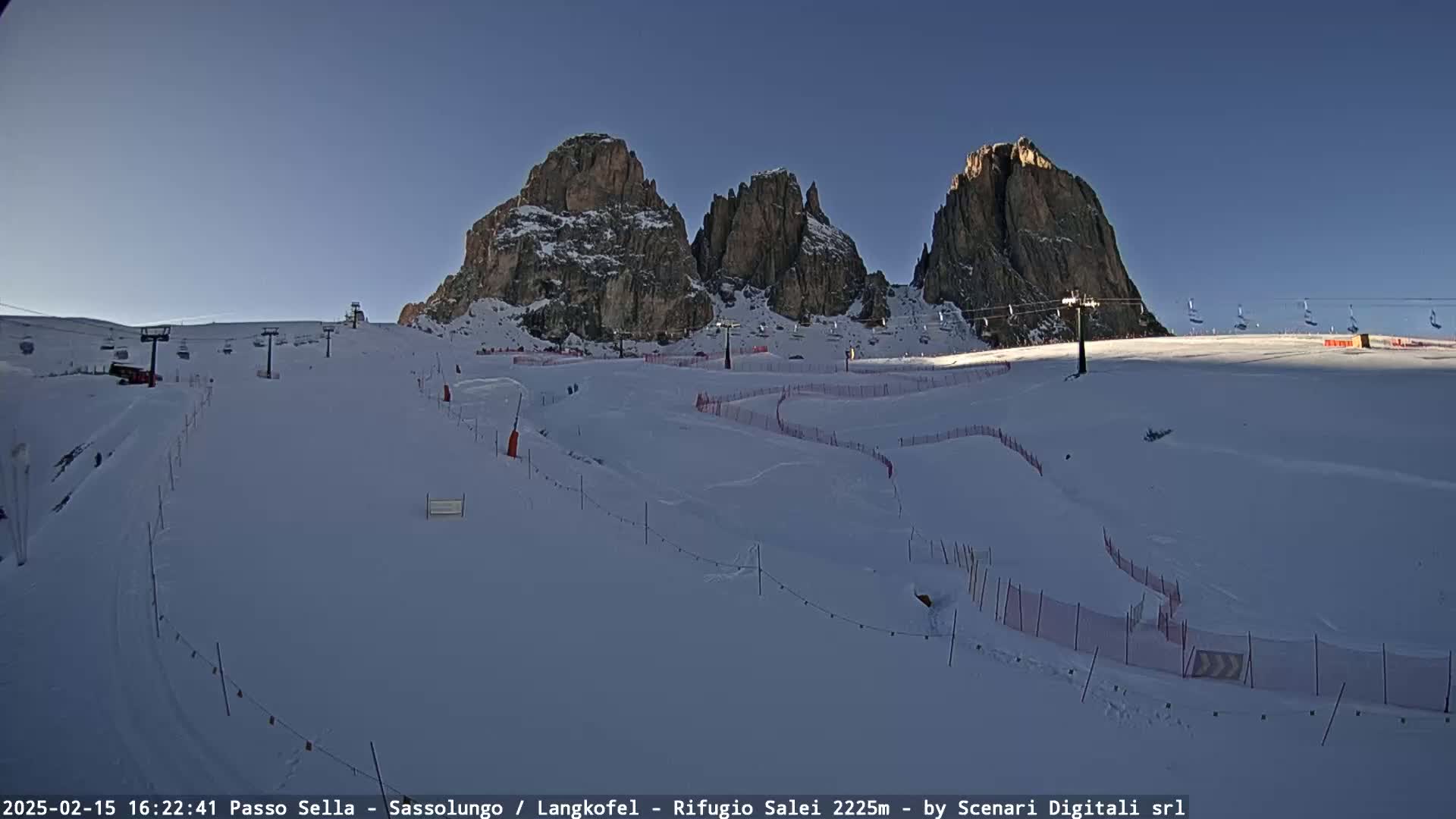 A snow-covered ski slope with chairlifts and tall rock formations in the background under a clear blue sky.