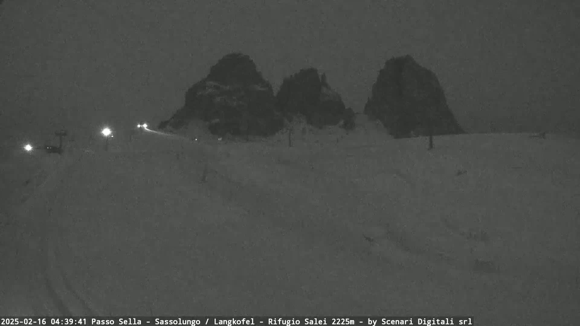 A snow-covered road with a few vehicles and lights leads toward three dark, snow-capped mountains under a nighttime sky.