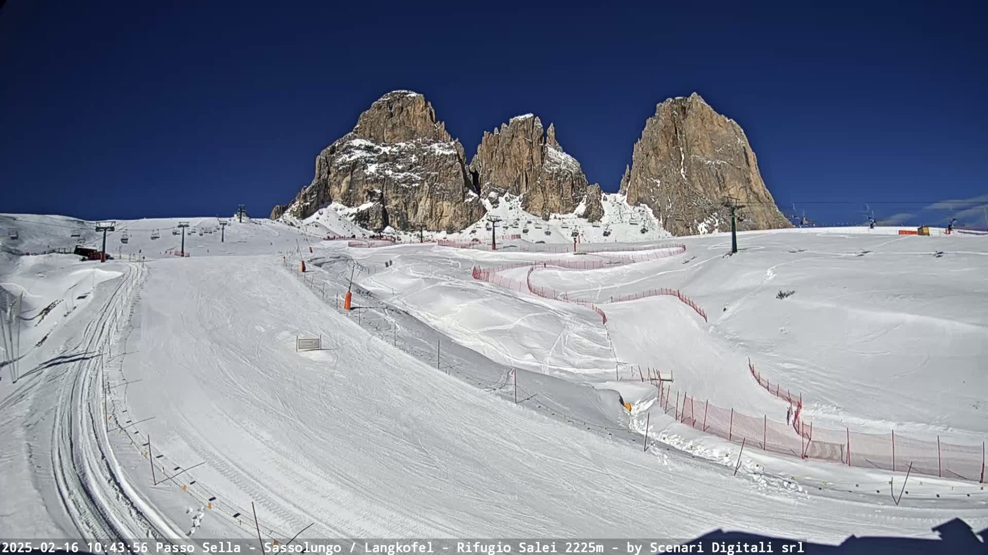 A snow-covered ski slope with several ski lifts and marked runs is set against a backdrop of jagged, snow-dusted mountains under a clear blue sky.