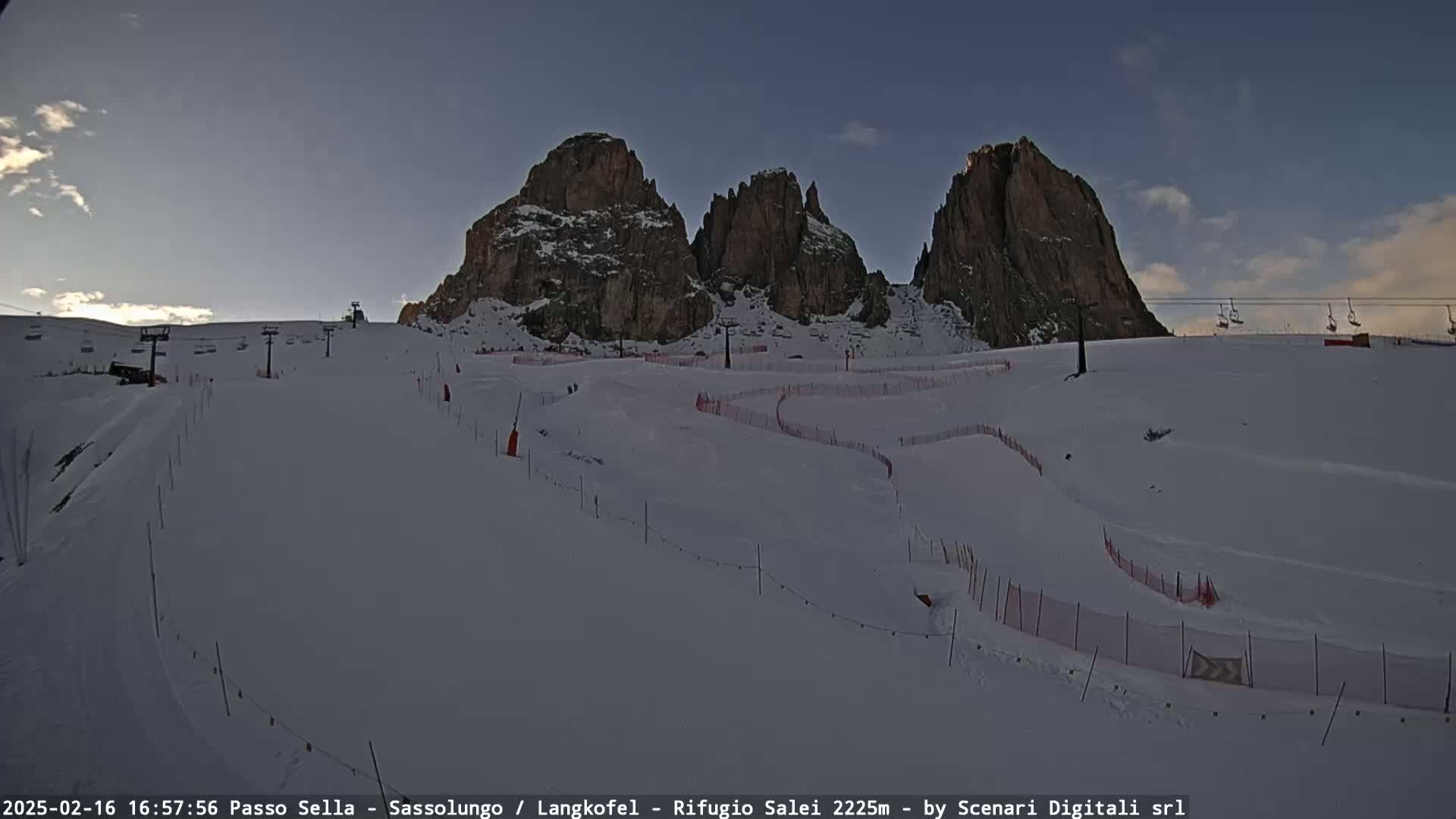 A snow-covered ski slope with several ski lifts and marked runs is situated below a range of rocky, snow-dusted mountains under a clear, twilight sky.