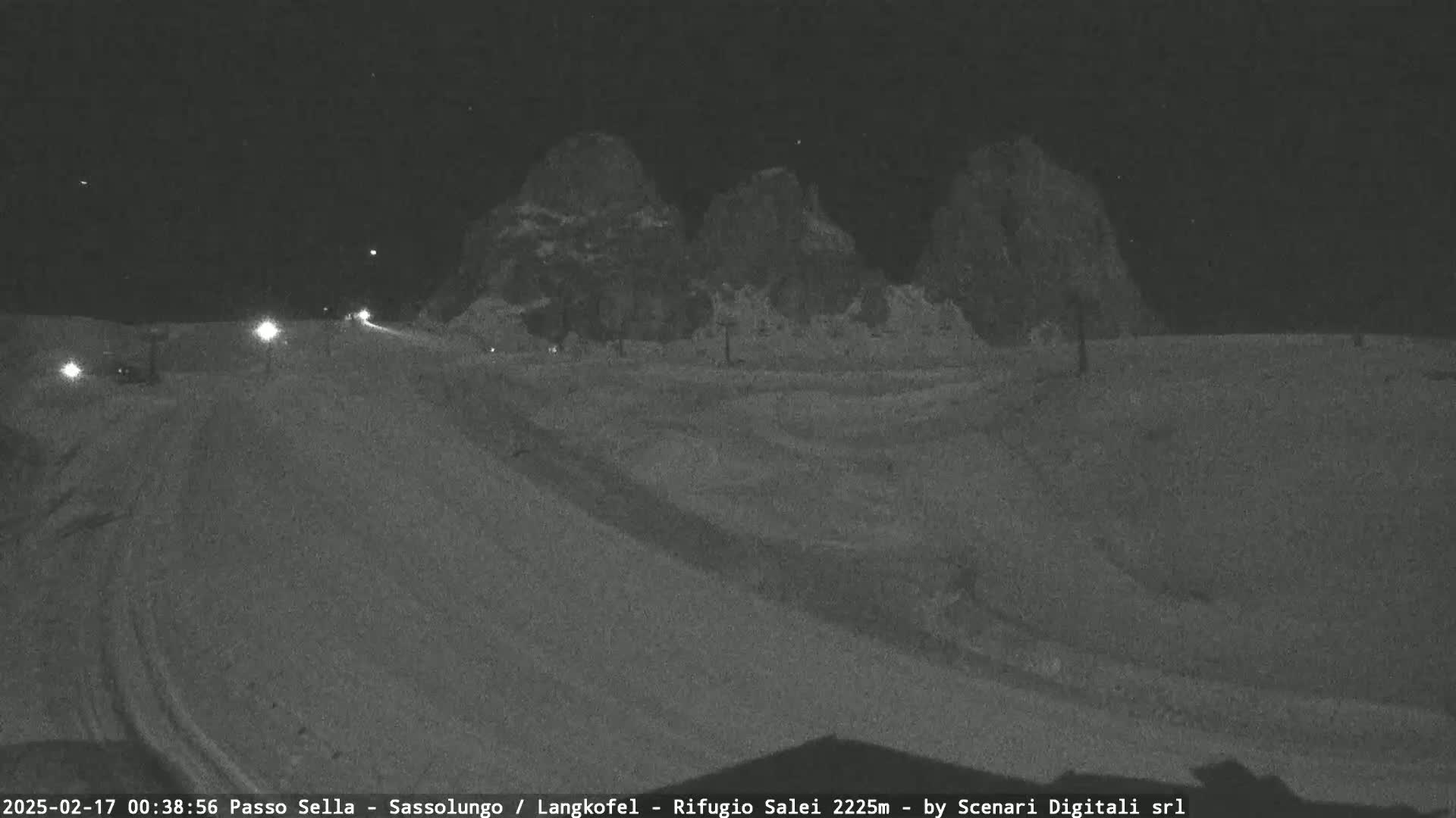 A snow-covered road at night is seen with a few vehicles driving towards three dark, rocky hills under a clear, dark sky.