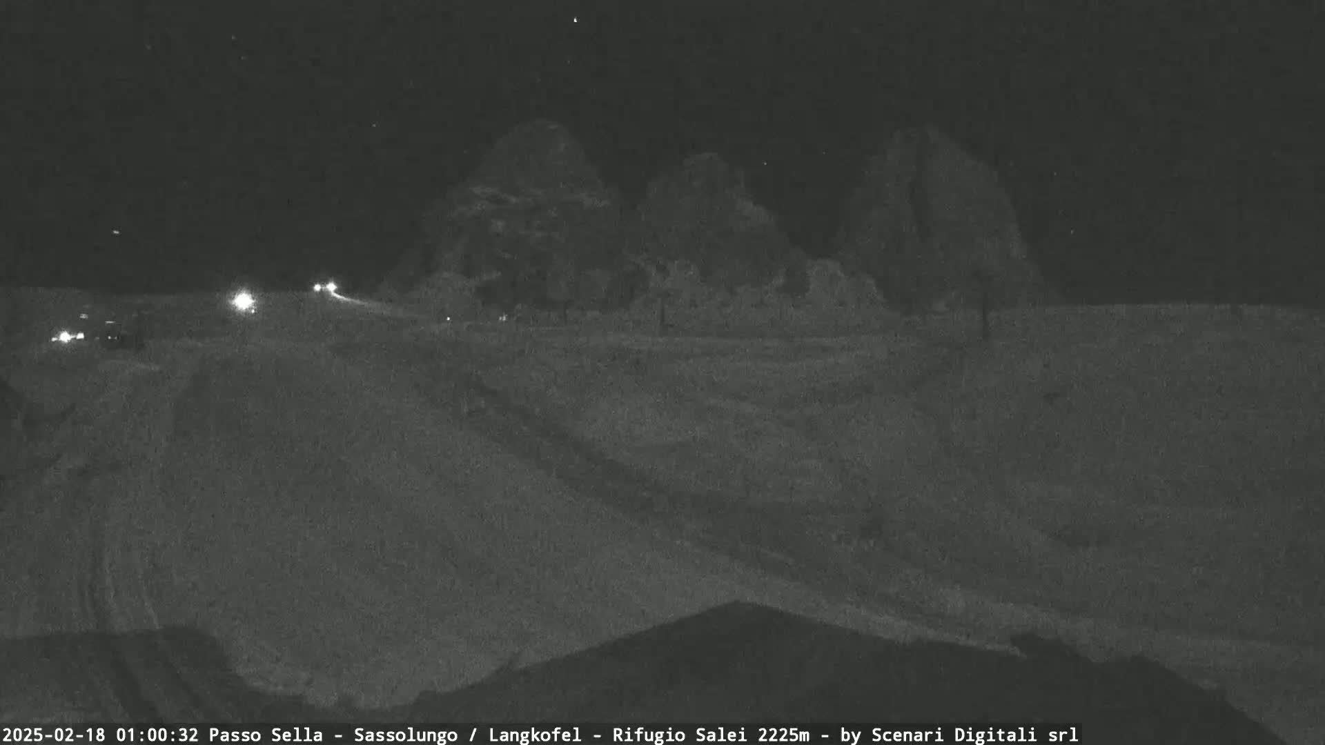 A nighttime, snow-covered road with a few vehicles driving towards dark, mountainous silhouettes under a clear sky.