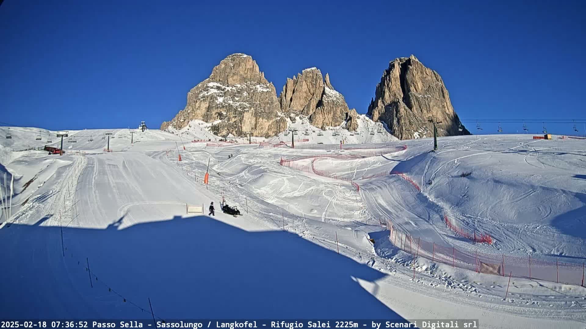 A snow-covered ski slope with several ski lifts and mountain peaks in the background under a clear blue sky.