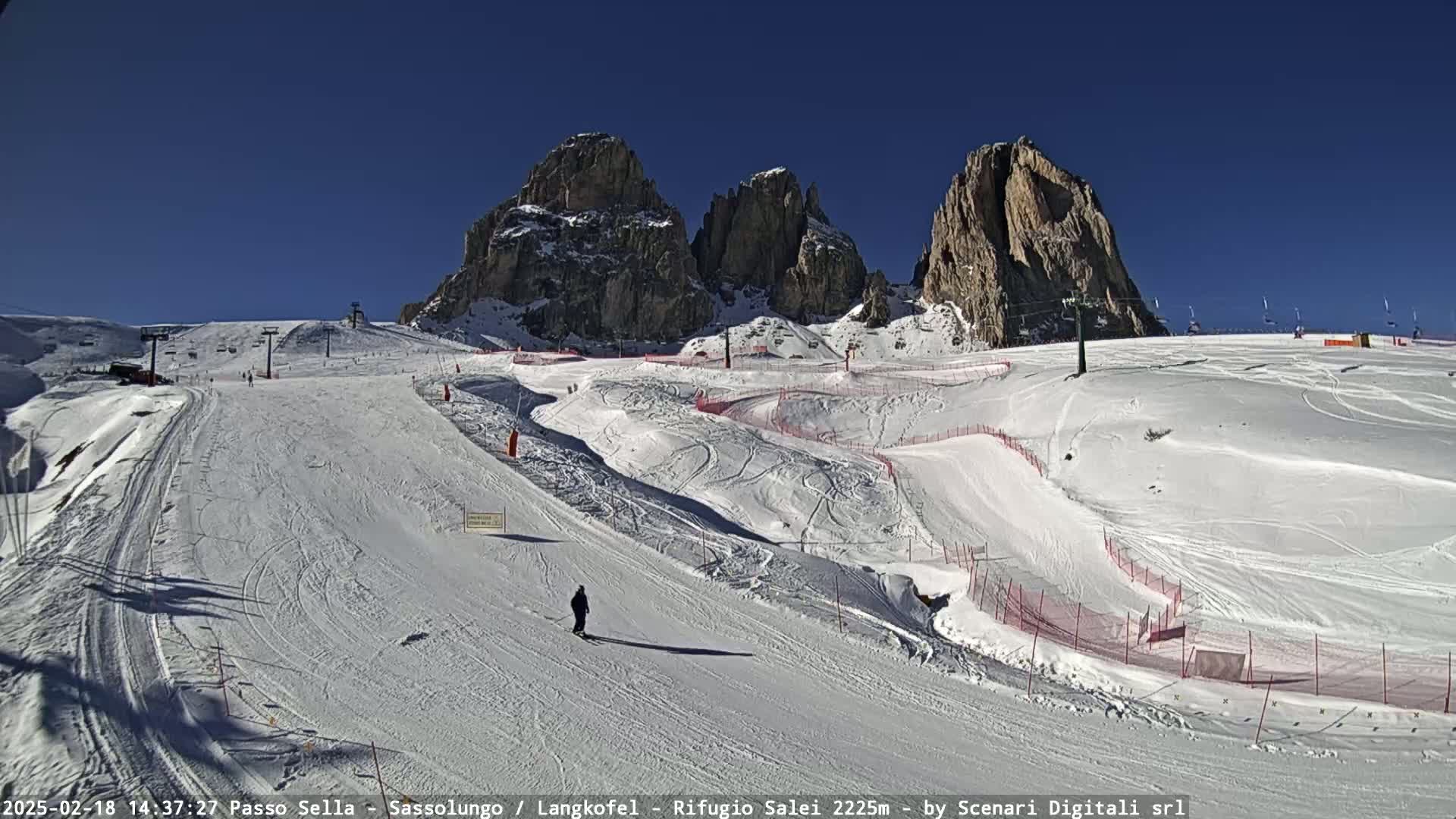 A snow-covered ski slope with several runs and a few skiers, situated below towering, rocky mountains under a clear blue sky.