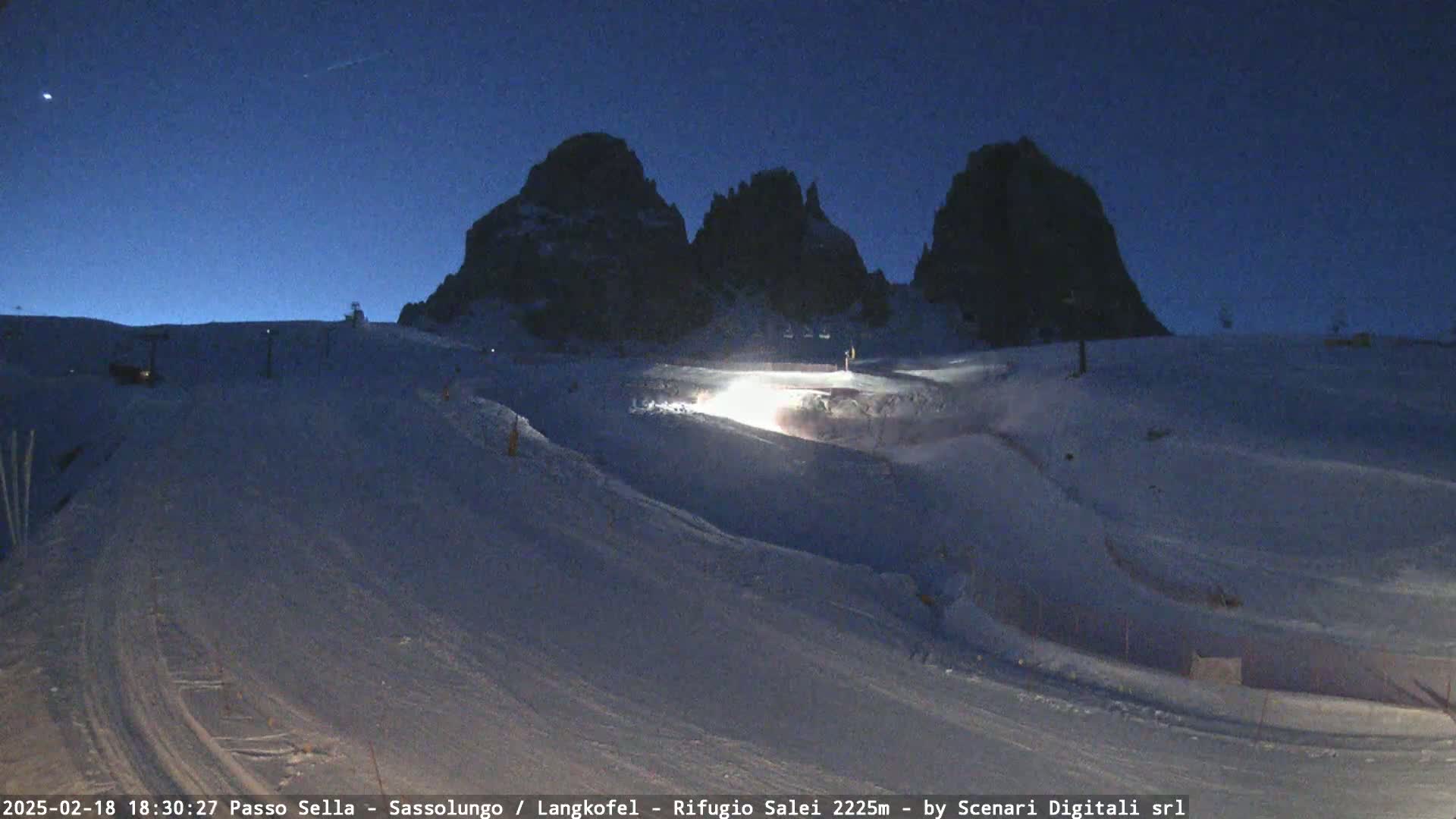 A snow-covered landscape at night, illuminated by a bright light source, with dark, rocky mountains in the background under a twilight sky.