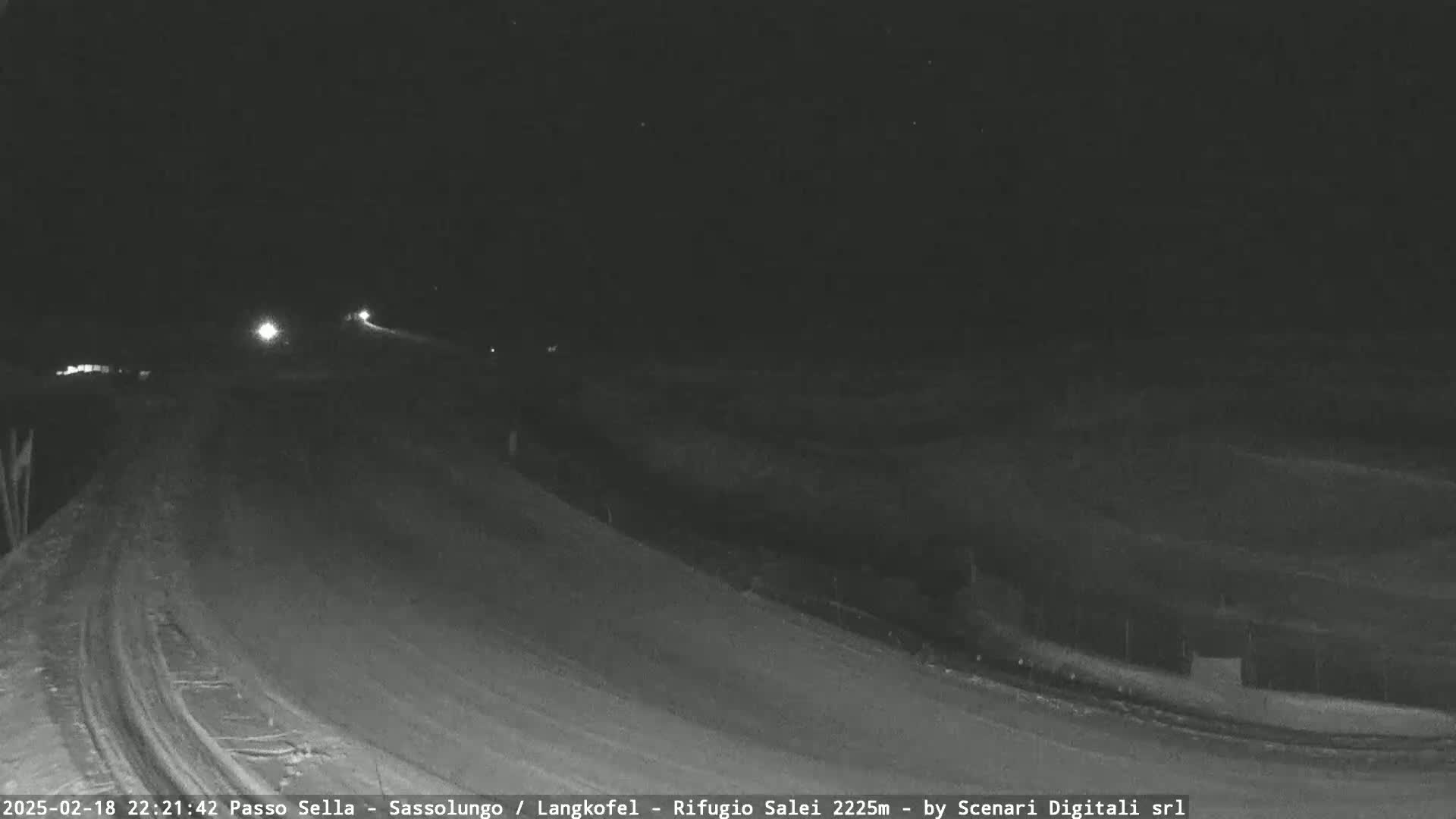 A snow-covered road curves gently to the left under a dark, nighttime sky, with a few vehicle headlights visible in the distance.