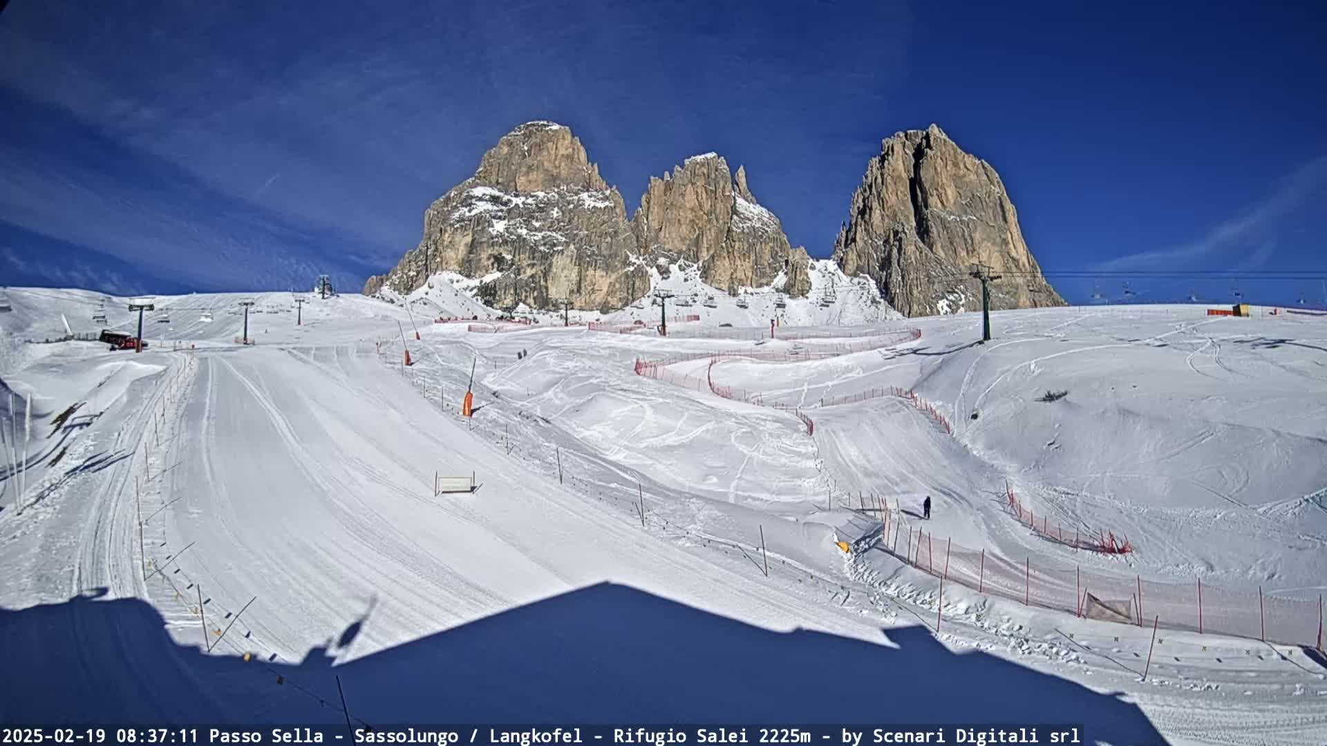 A snow-covered ski slope with several ski lifts and rocky mountain peaks in the background under a clear blue sky.